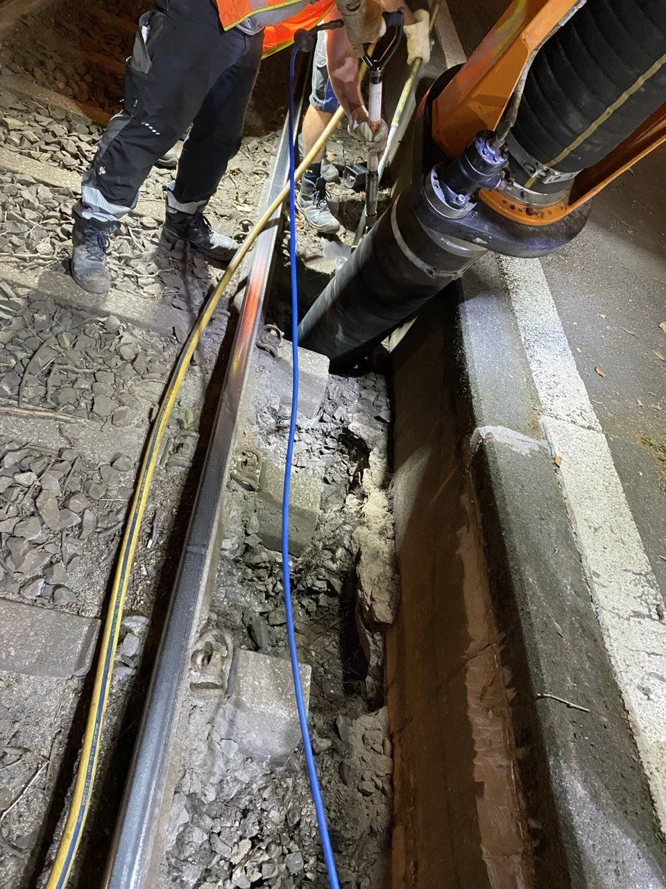 Arbeitskräfte beim Gleisbau mit einem großen Bohrer und Werkzeug auf einer Baustelle neben der Straße.