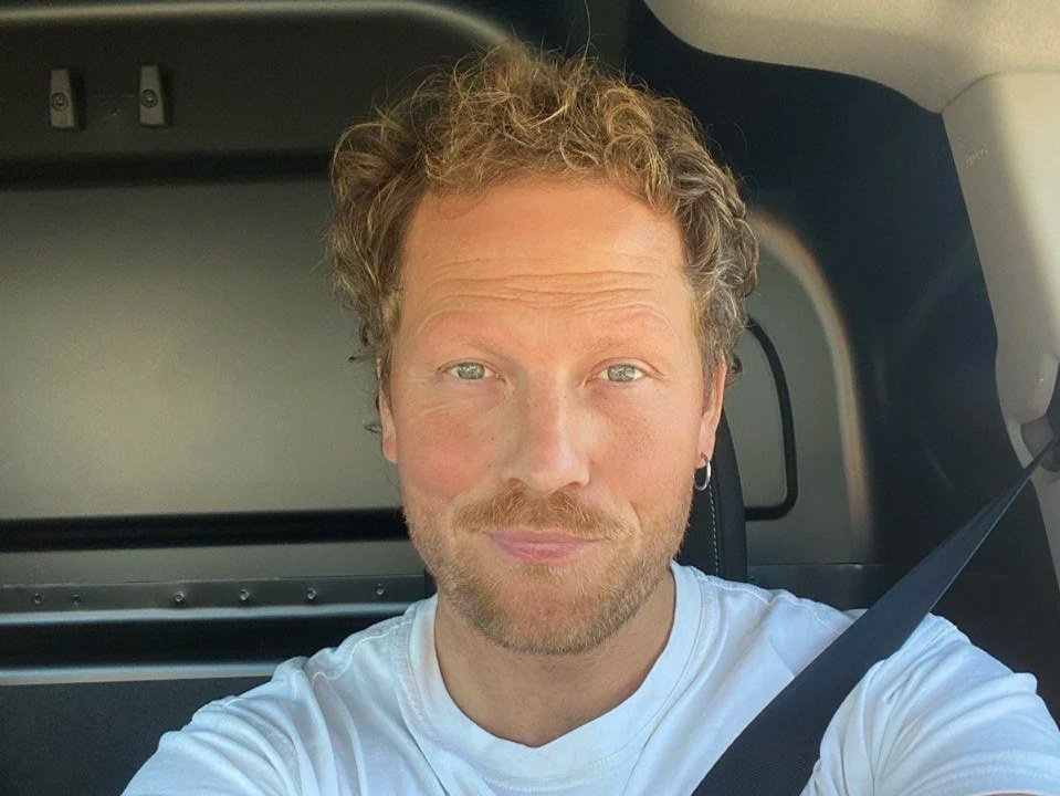 Ein Mann mit lockigem, braunem Haar und einem kurzen Bart trägt ein weißes T-Shirt und sitzt im Auto, trägt einen Sicherheitsgurt.