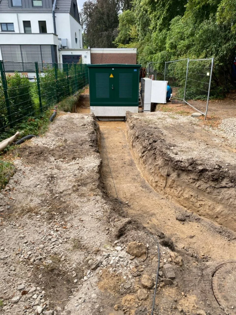 Baustelle mit Grube und verlegte Kabel, Zaun und eine kleine Gebäude, Bäume im Hintergrund.