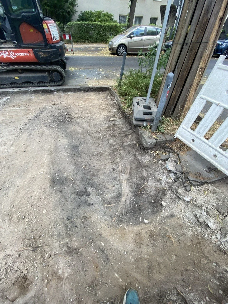 Baustelle mit aufgebrochener Erdoberfläche, Baumaschine und Bauzaun, vor Häusern und parkenden Autos.