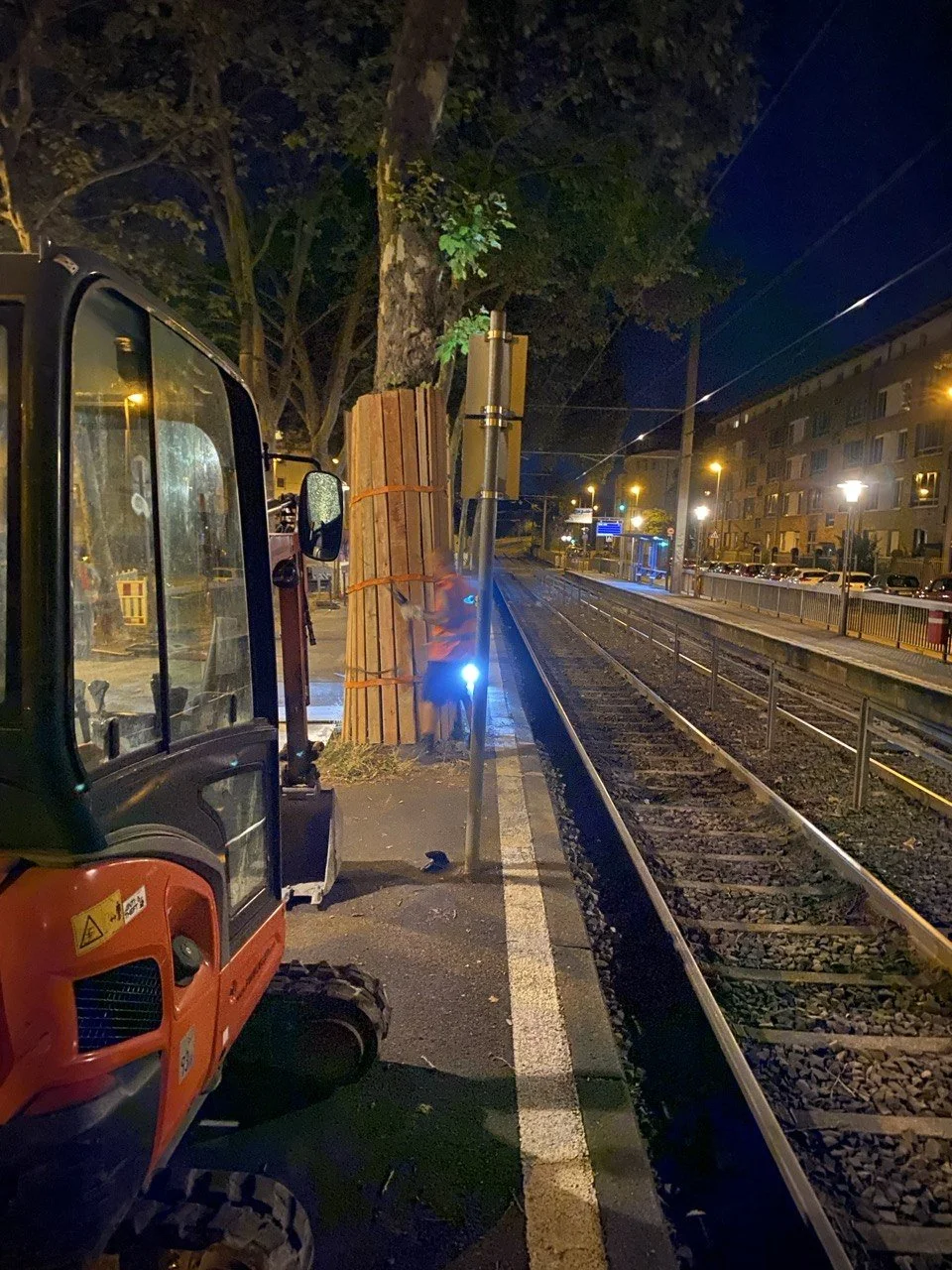 Bauarbeiten an einem Bahnsteig bei Nacht, mit Baufahrzeug, Holzstämmen und einem Arbeiter, der eine Sicherheitsweste trägt.