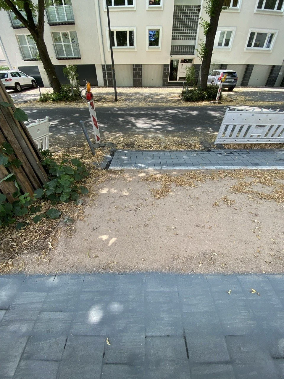 Straße vor einem modernen Mehrfamilienhaus mit Parkplätzen, Bäumen und Bauarbeiten, Absperrungen und Baumaschinen