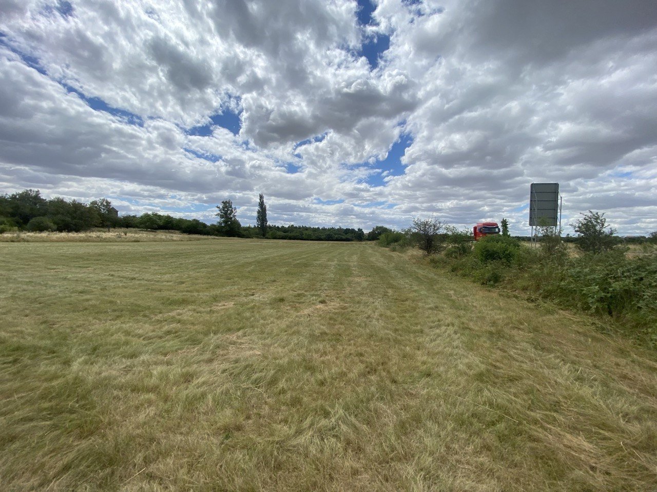 Ein offener, grüner Feldweg unter einem bewölkten Himmel, mit Büschen und einem roten Fahrzeug am Straßenrand.