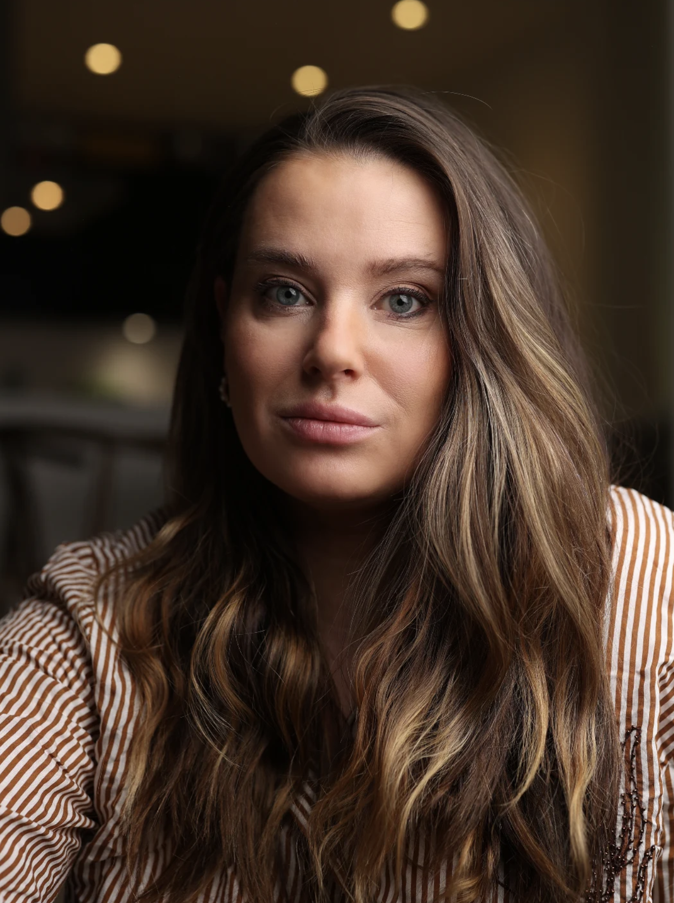 Nuorekas nainen, keskellä kuva, pitkät ruskeat hiukset ja vaaleansiniset silmät, pukeutunut raidalliseen vaatteeseen, istuu sisätiloissa.