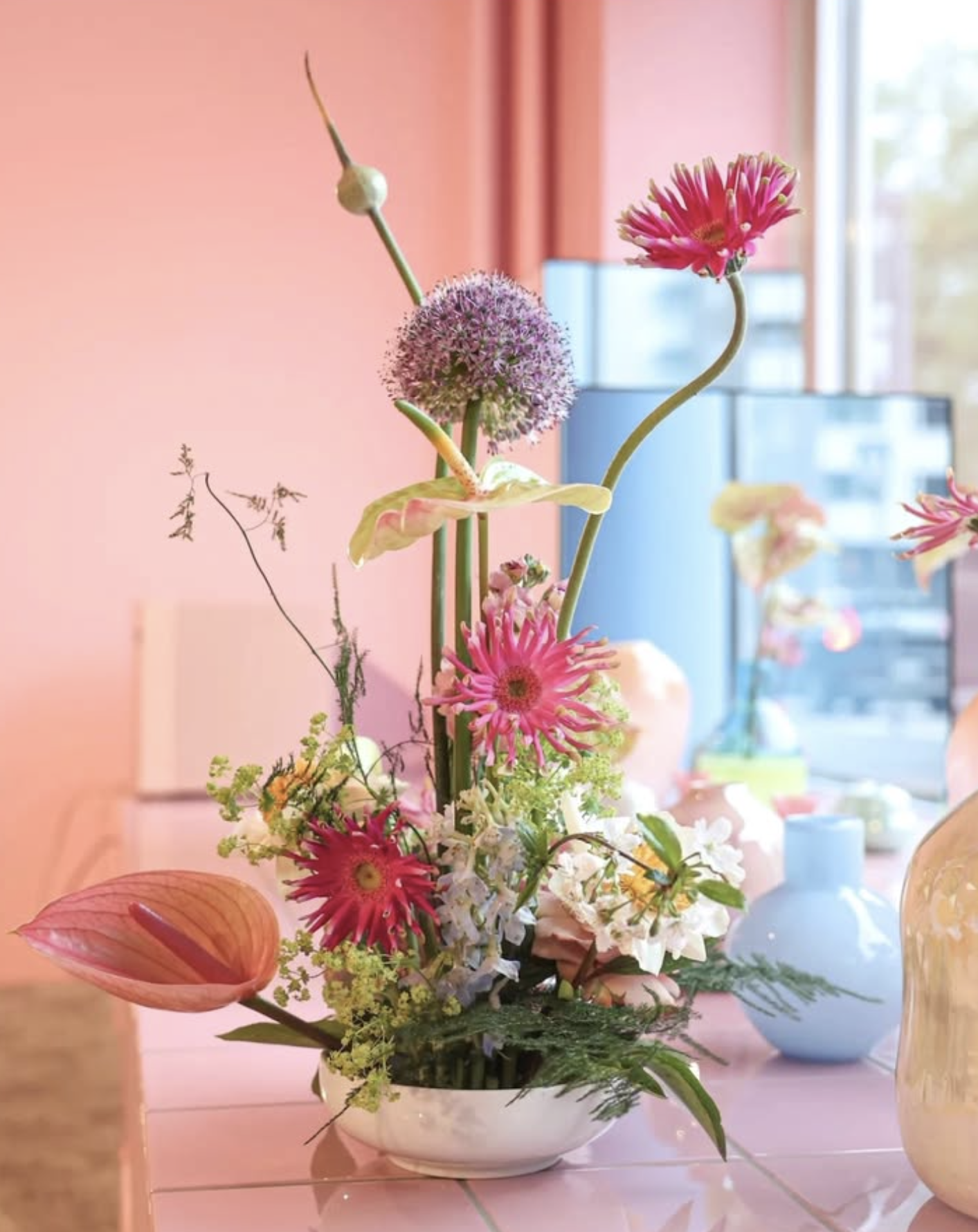 Värikäs asetelma kukkia vaaleanpunaisella pöydällä ikkunan edessä.