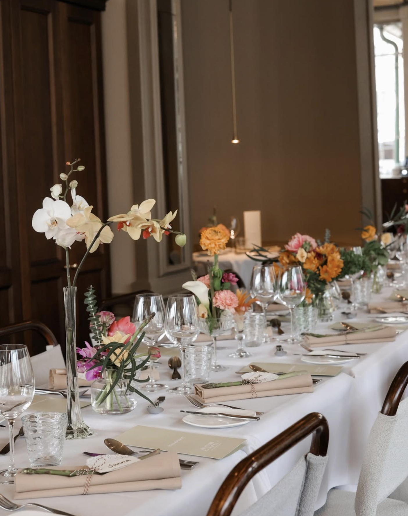 Pitkä juhlapöytä koristeltuna kukka-asetelmilla, laseilla, lautasilla ja tietokortteilla, sisätiloissa.