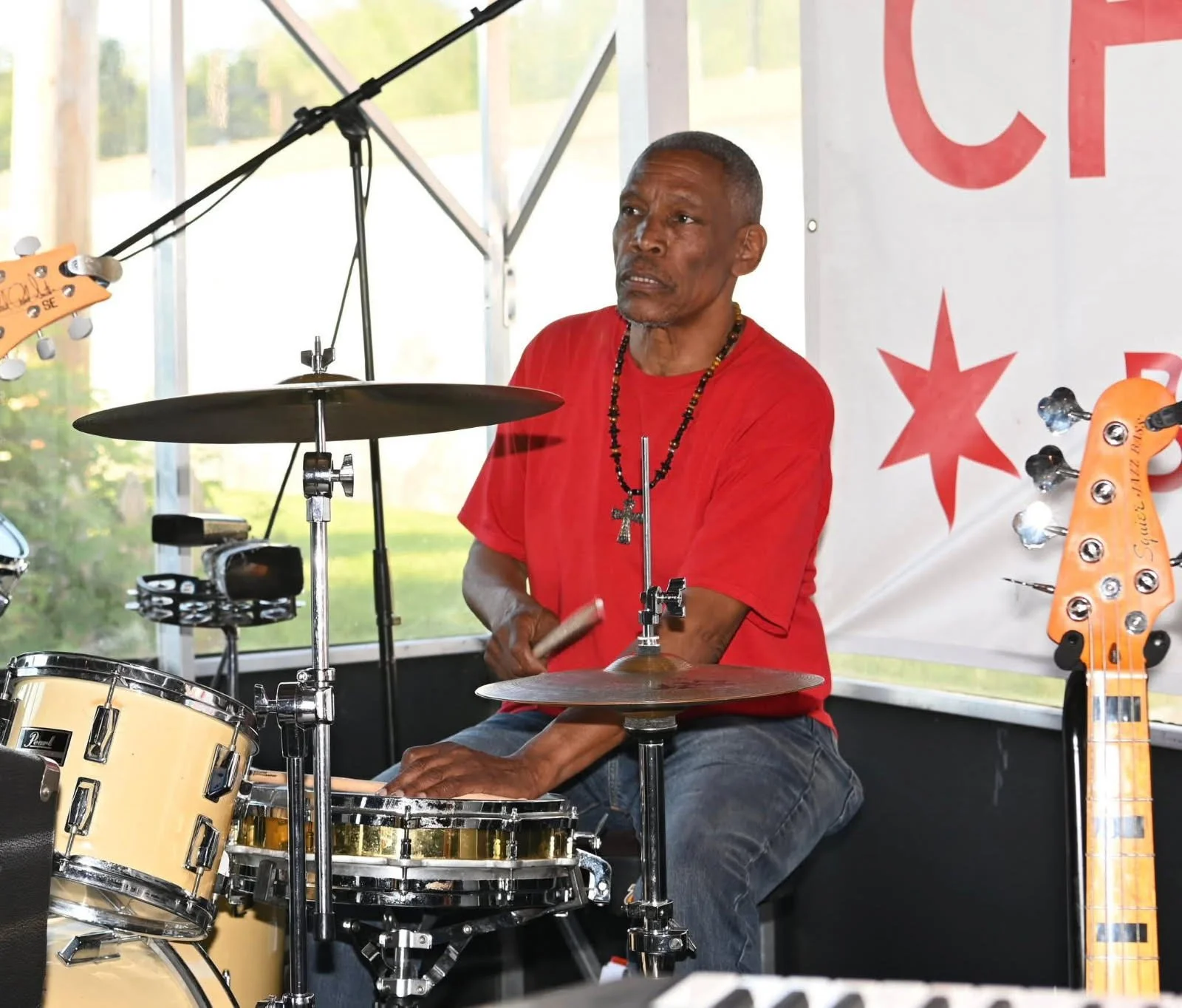 A man playing a drum set on stage, wearing a red shirt and necklace with a cross, with a bass guitar visible on the right and a large banner with red star symbols in the background.