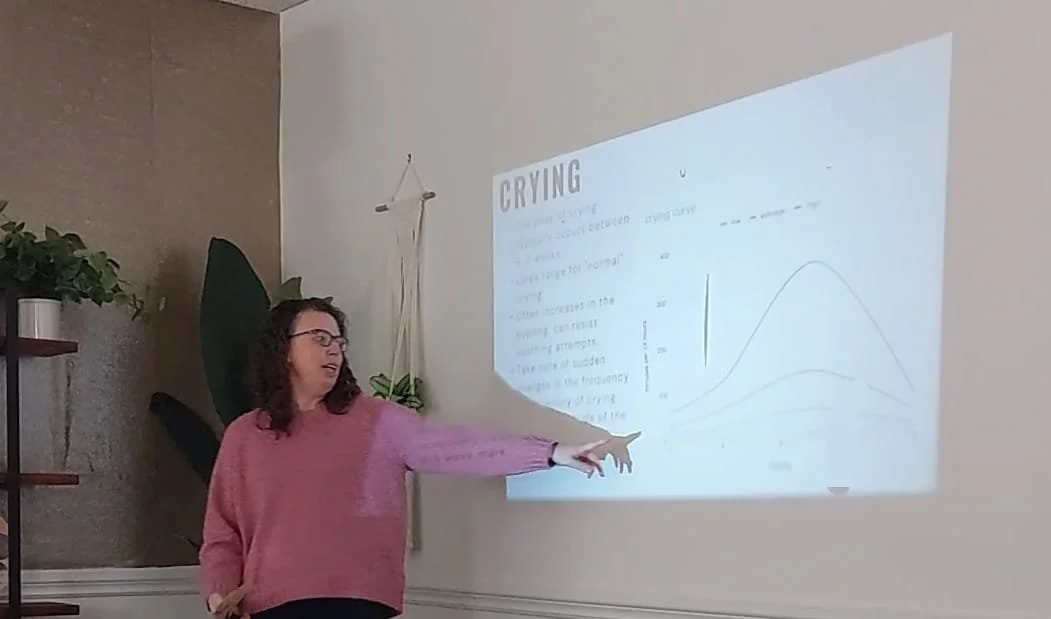 Kelly pointing to a presentation slide about infant crying patterns during a newborn care class.