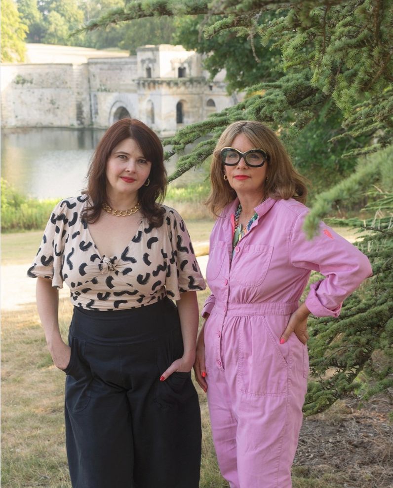 Two women standing outdoors next to a pine tree, with a historic stone bridge and water in the background. One woman is wearing a leopard print blouse and dark pants, the other is wearing a pink jumpsuit and sunglasses.