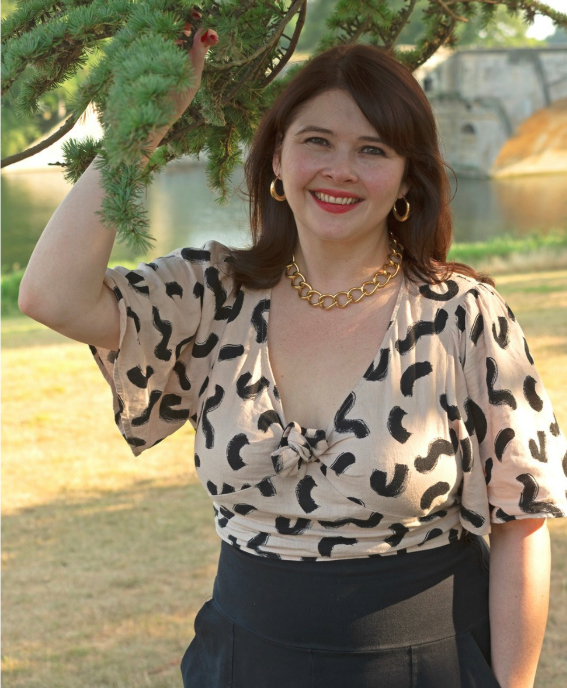 A woman with auburn hair, wearing a leopard print blouse, gold jewelry, and black pants, stands outdoors near a tree, smiling at the camera.
