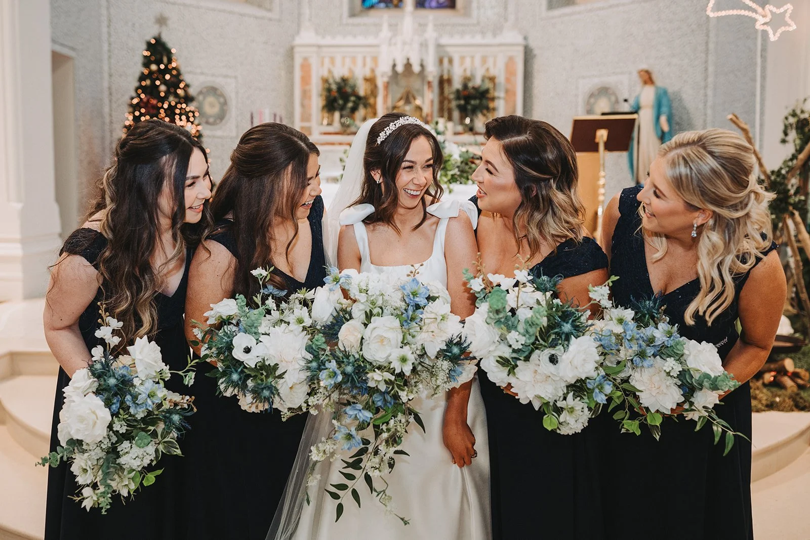 A bride and her four bridesmaids smiling and looking at each other, holding large floral bouquets, inside a decorated church.