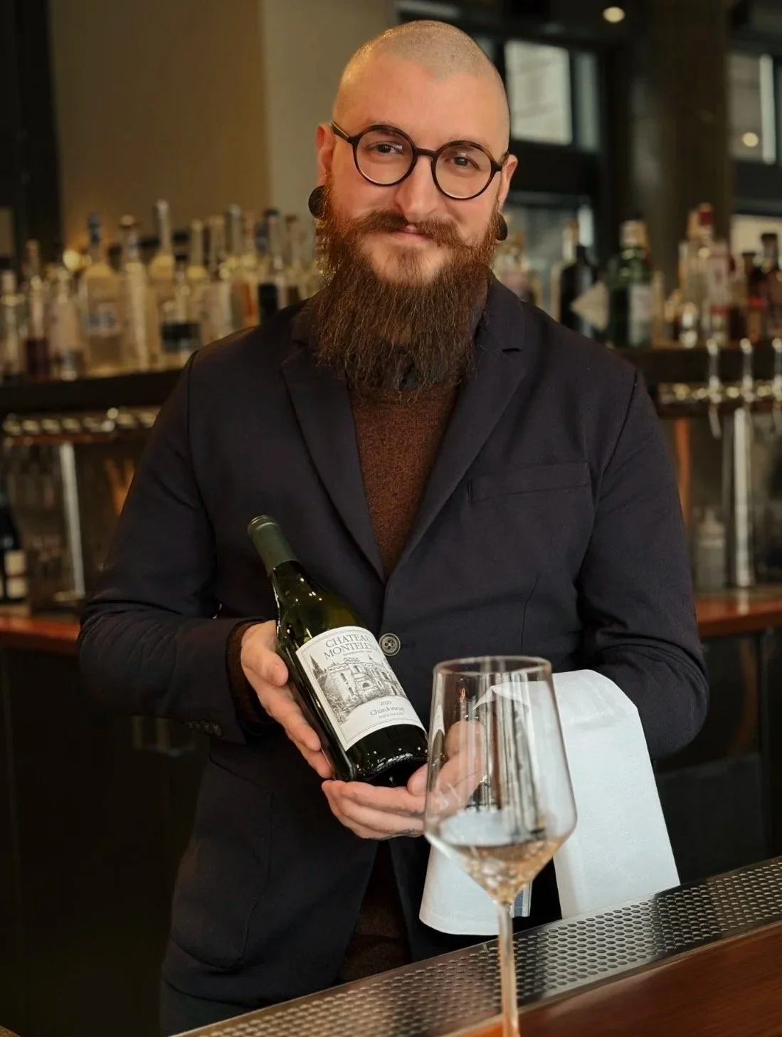 A man with glasses and a beard holding a bottle of red wine in a restaurant or bar setting
