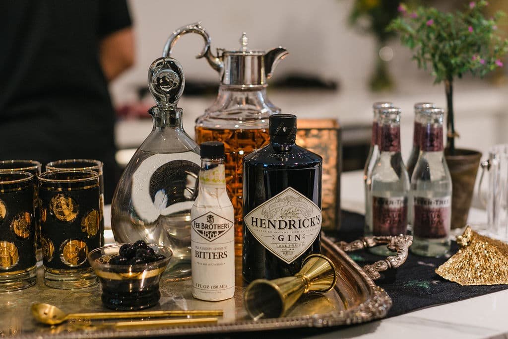 A silver tray with bottles of Hendrick's gin, Fee Brothers bitters, a glass decanter, and various glasses, set on a black surface with a potted plant in the background.