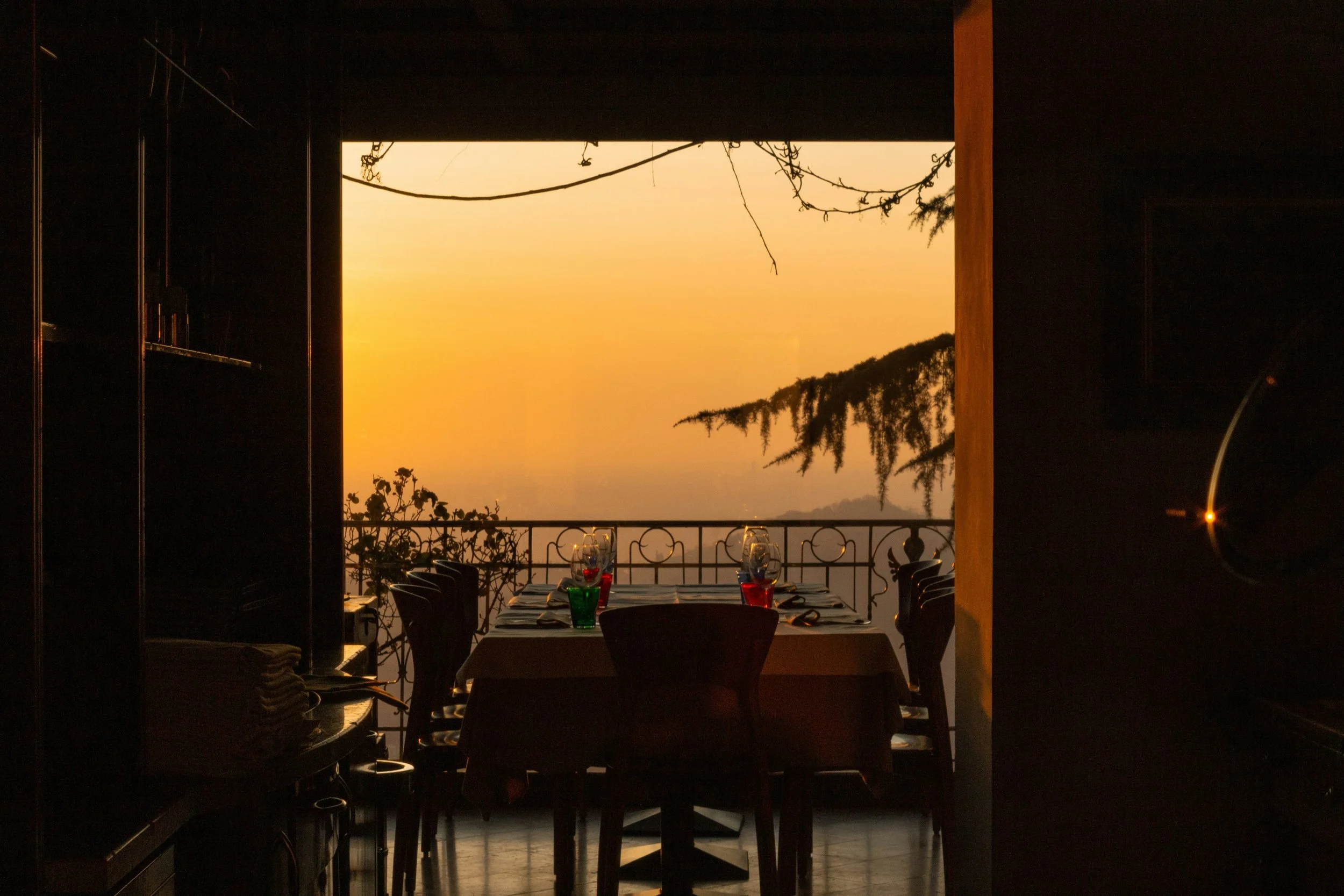 Indoor dining area with a large open window showing a sunset view, tables set with glasses and napkins, chairs around the tables, and decorative plants near the window.