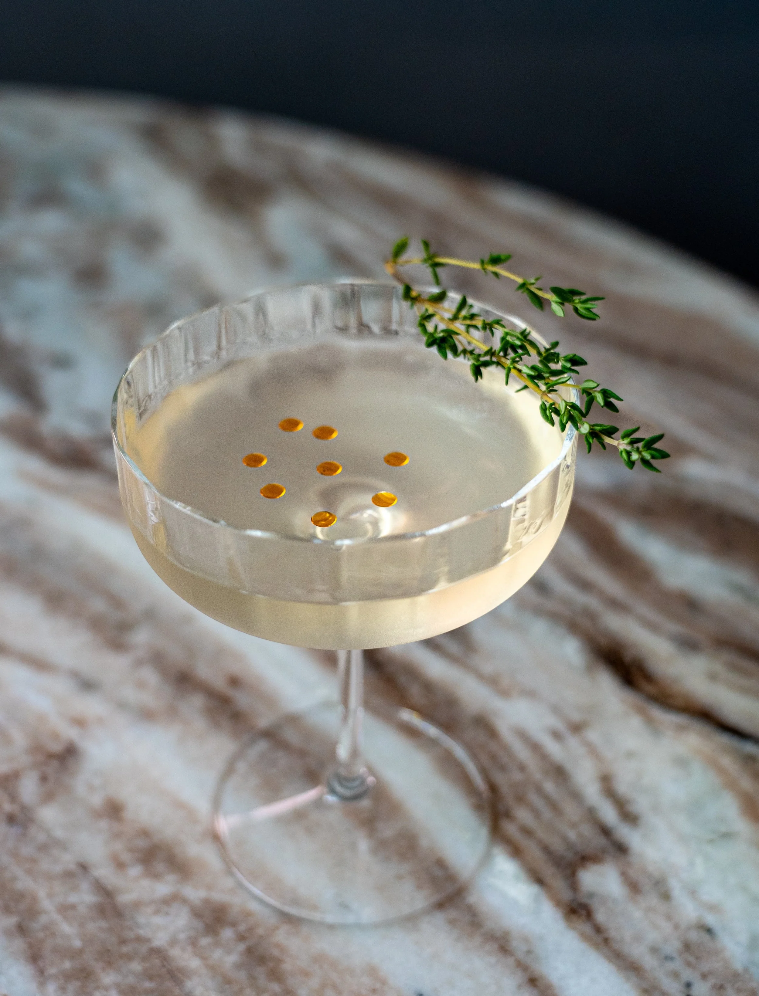 A cocktail in a coupe glass garnished with a sprig of thyme, with some orange round edible decorations floating on top, placed on a marble surface.