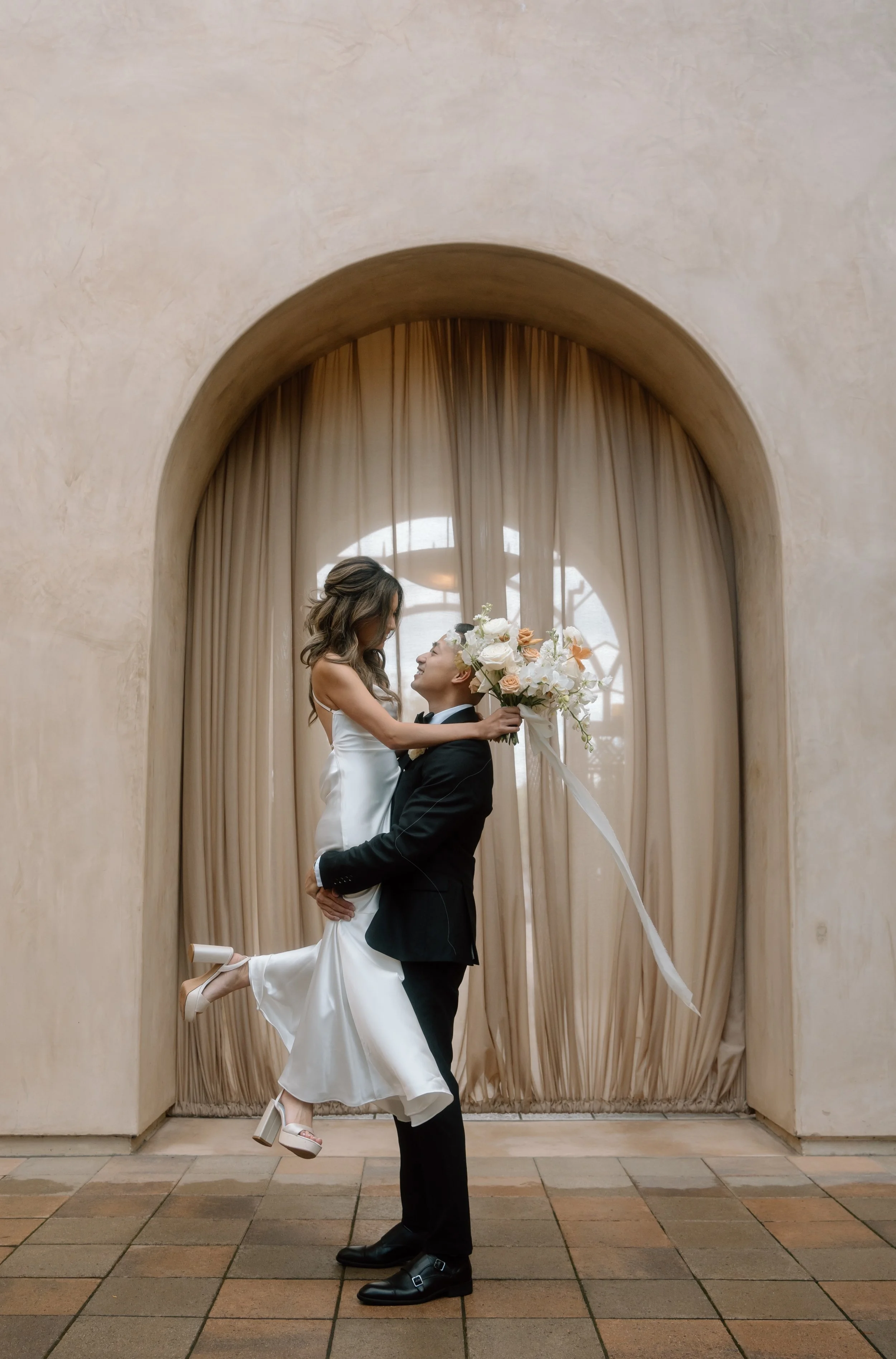 Bride and groom in embrace, groom lifting bride, bride holding bouquet, in front of arched backdrop with curtains.