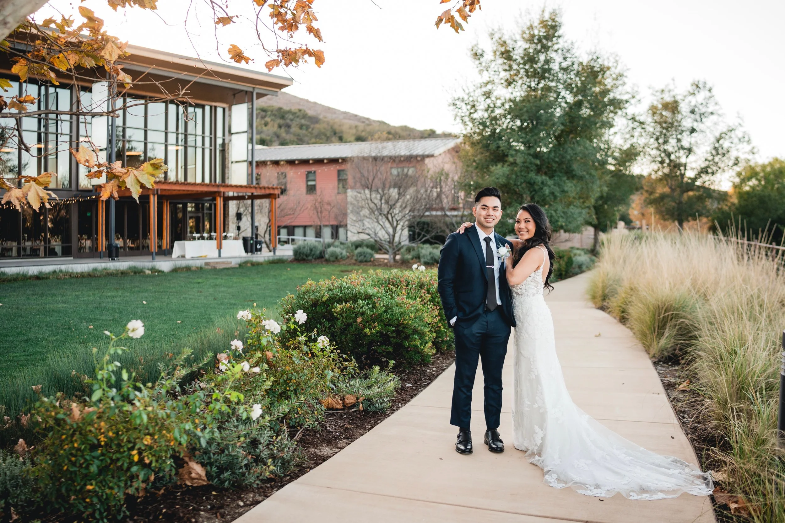 A couple in formal attire posing outdoors, standing on a pathway next to landscaped gardens, with a modern building in the background. The woman is wearing a white dress and the man is in a dark suit.