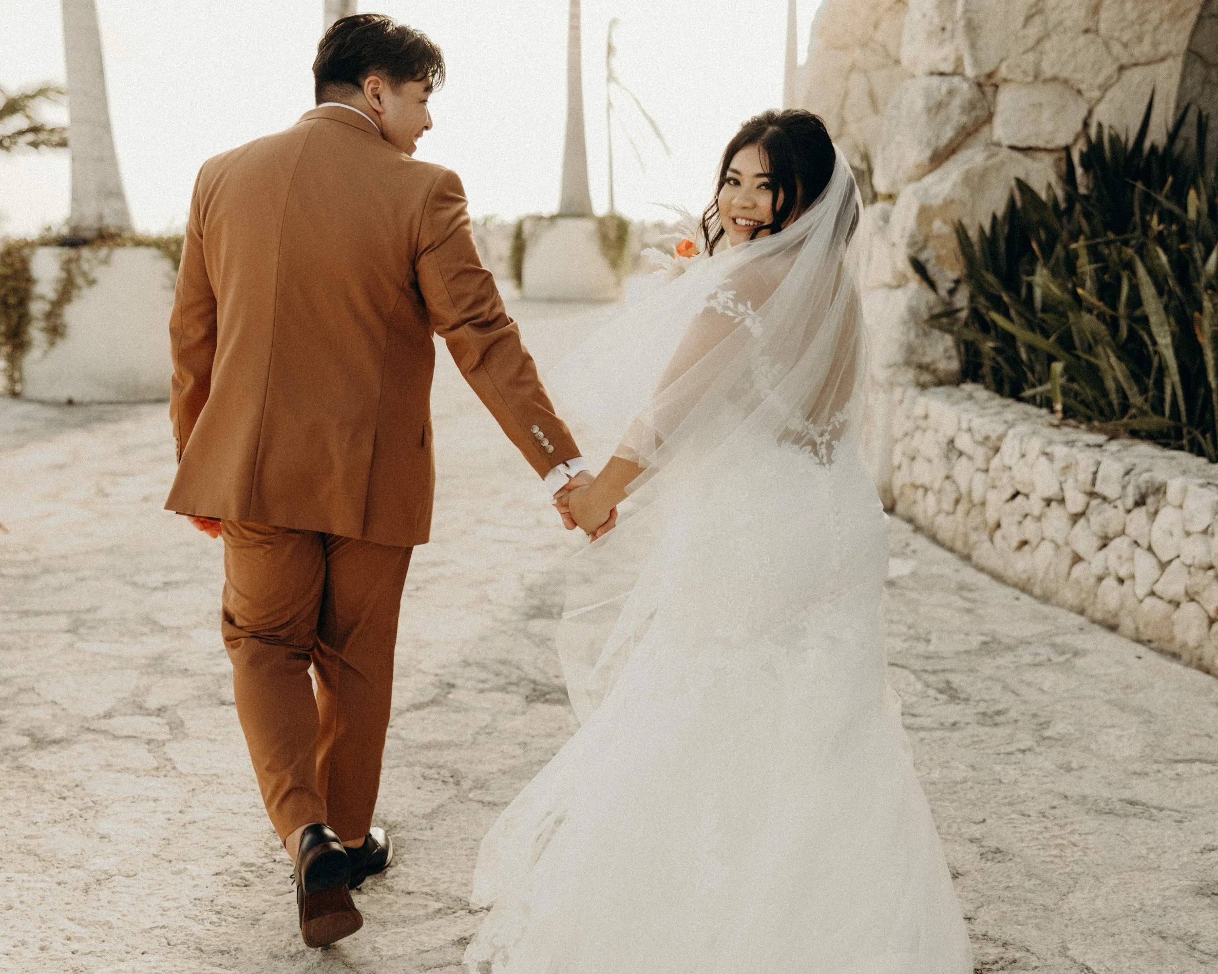 Bride and groom walking hand in hand on a stone path, bride wearing a white dress and veil, groom in a brown suit, with plants and stone wall in the background.