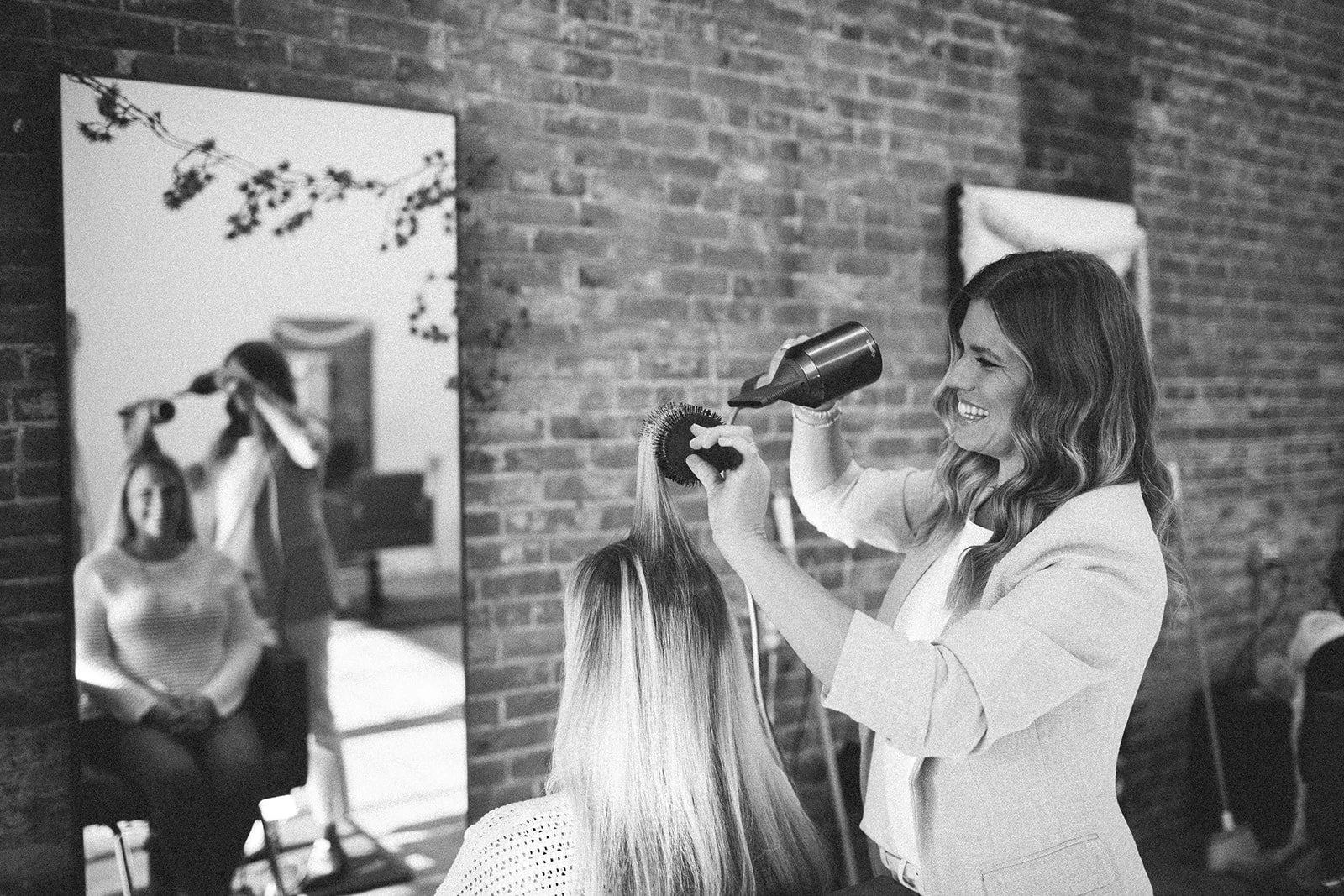 A woman styling another woman's hair in a salon with exposed brick walls, using a hairdryer and brush, while a mirror reflects the scene.