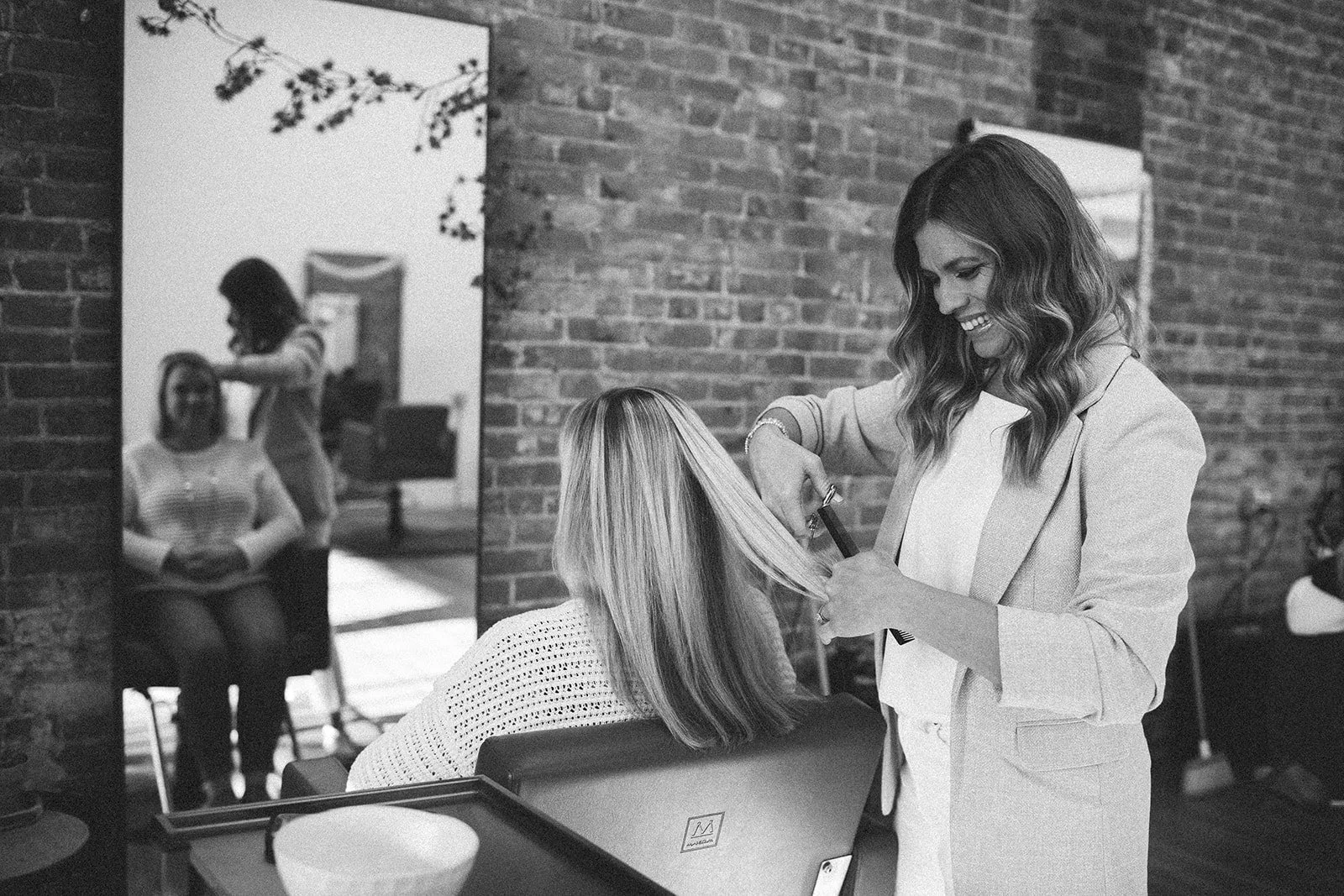 A hairstylist cutting a woman's hair in a salon with exposed brick walls, seen through a large mirror.