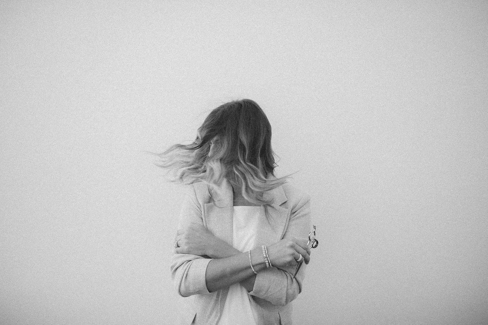 Black and white photo of a woman with shoulder-length hair turned away, holding scissors and a comb, standing against a plain background.