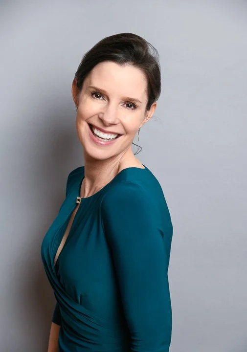 A woman with short dark hair smiling against a light gray background, wearing a teal dress.