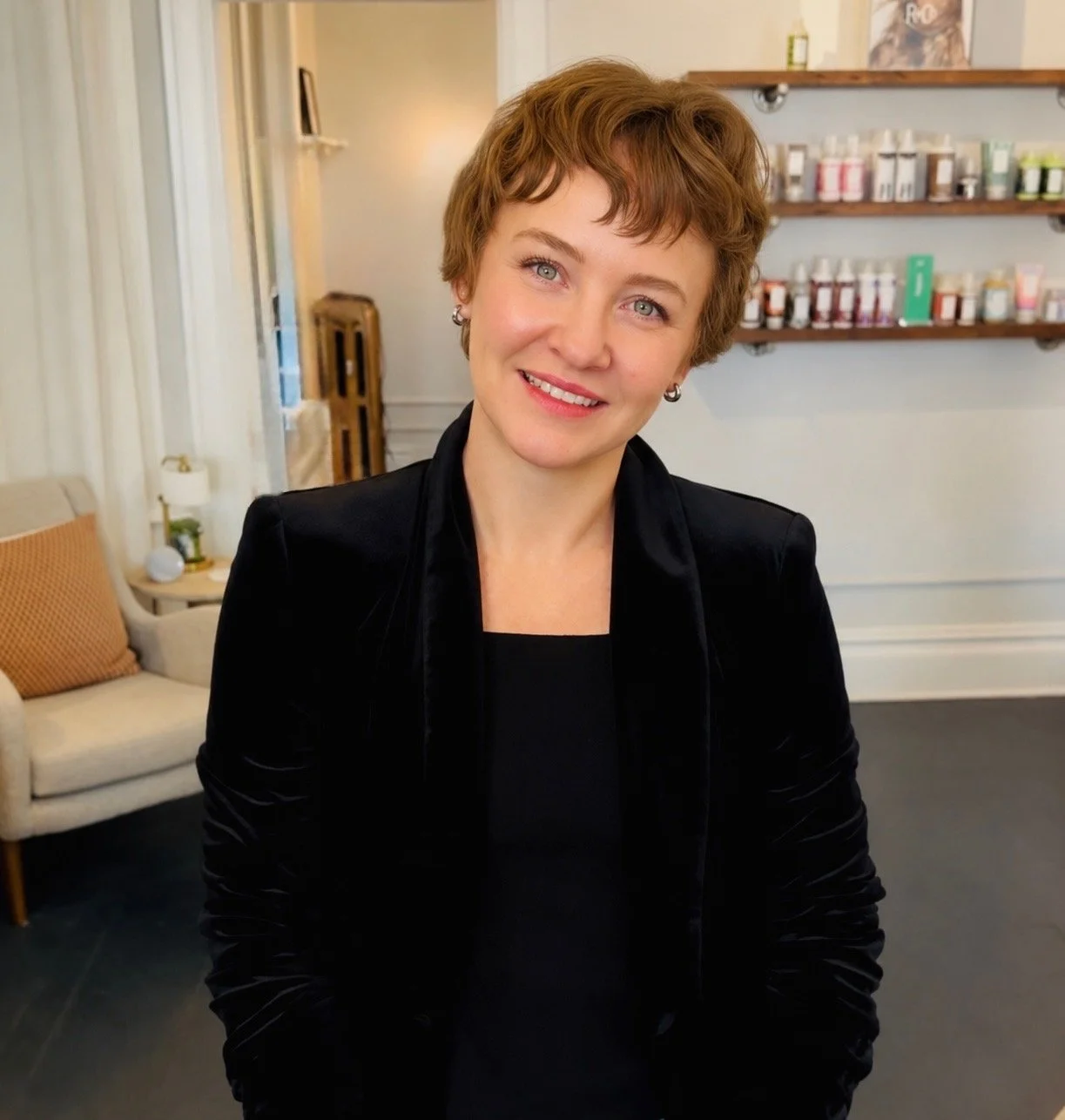 A woman with short, curly red hair and blue eyes, wearing a black blazer, smiling in a cozy, well-lit room with a beige couch, some cushions, and shelves filled with small bottles or containers in the background.