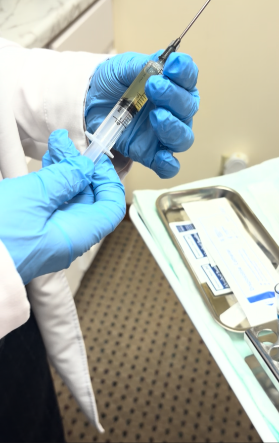 A healthcare worker wearing blue gloves handles a syringe, preparing an injection, in a medical setting.