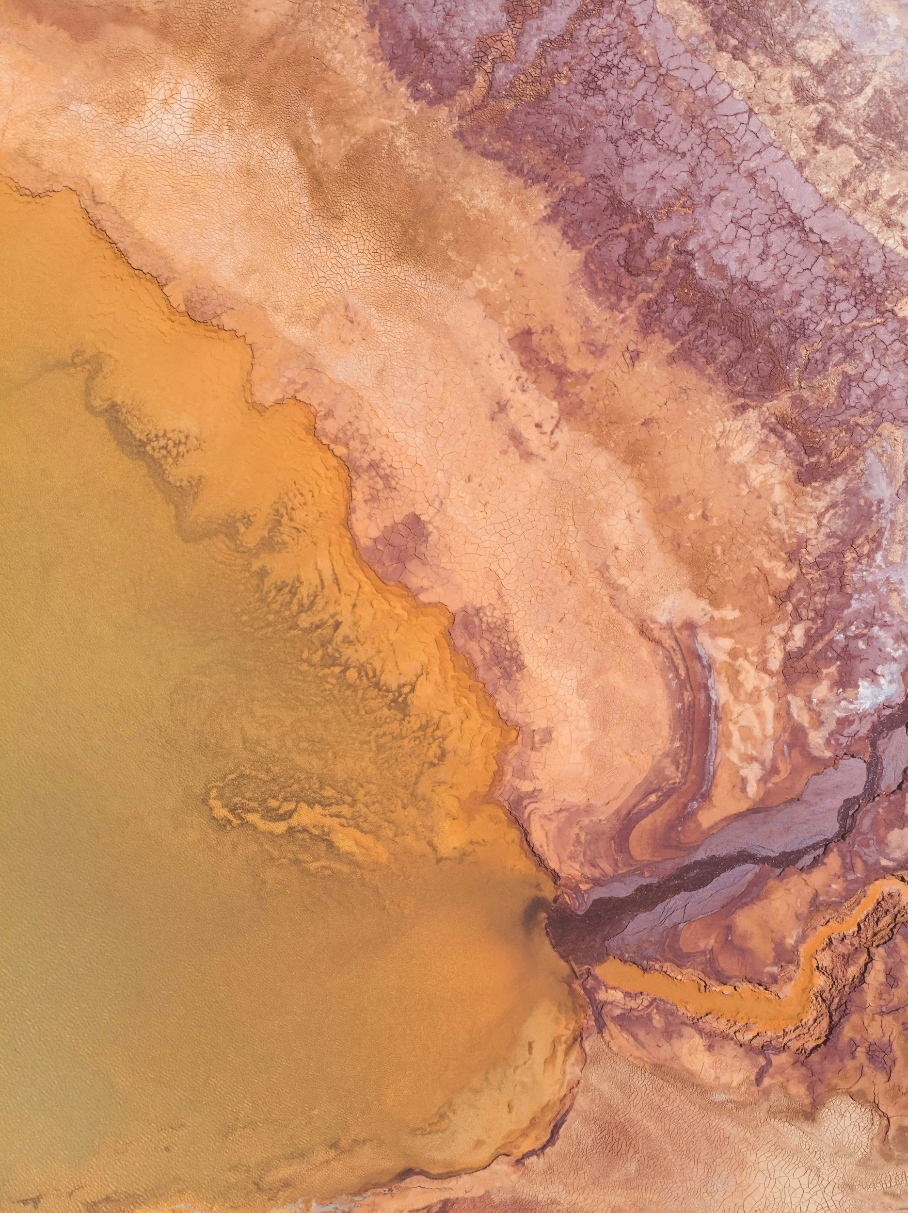 Aerial view of a desert landscape with various shades of brown, purple, and yellow, featuring cracked dry earth and winding formations.