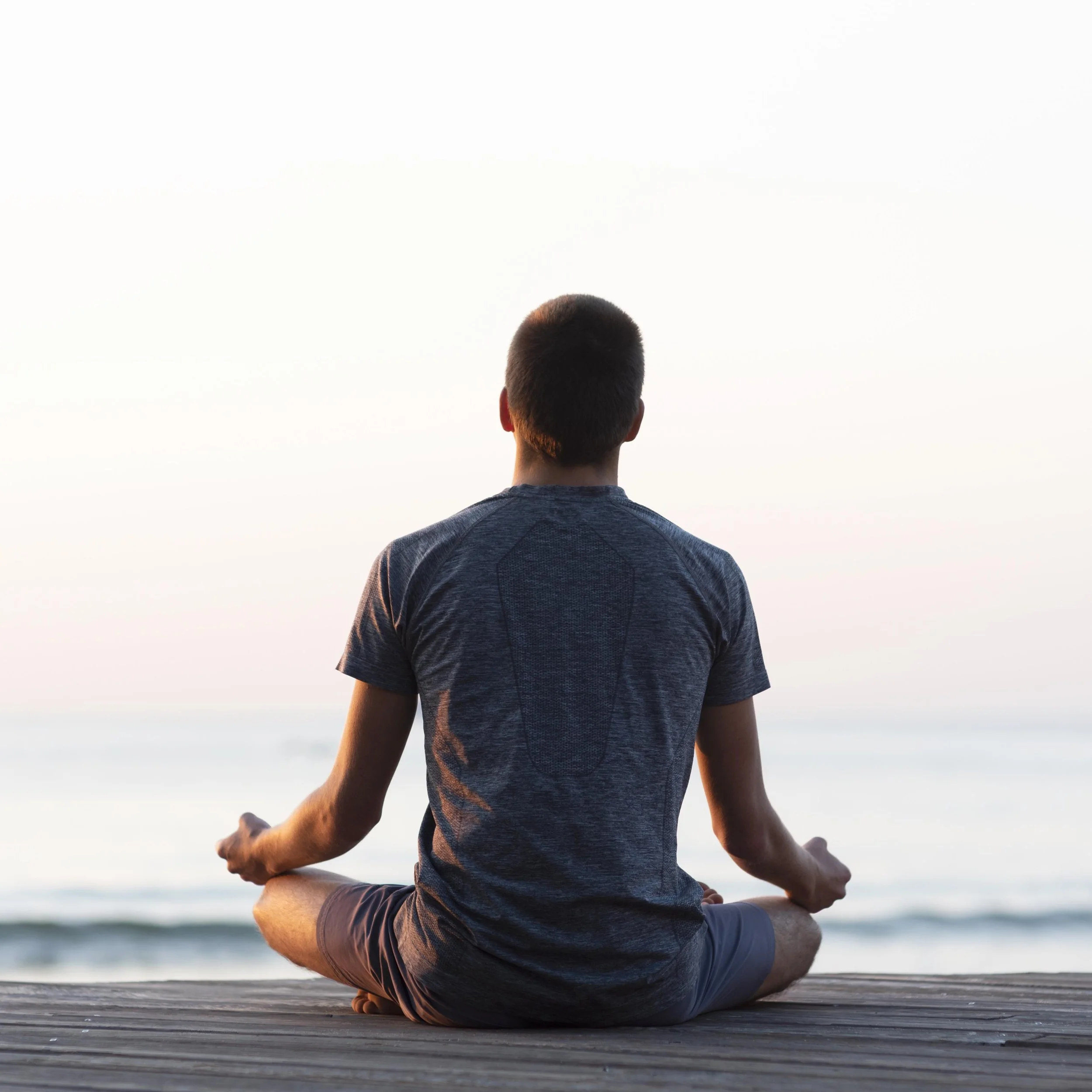 A person practicing yoga outdoors on a wooden deck, sitting cross-legged and facing the ocean during sunset.