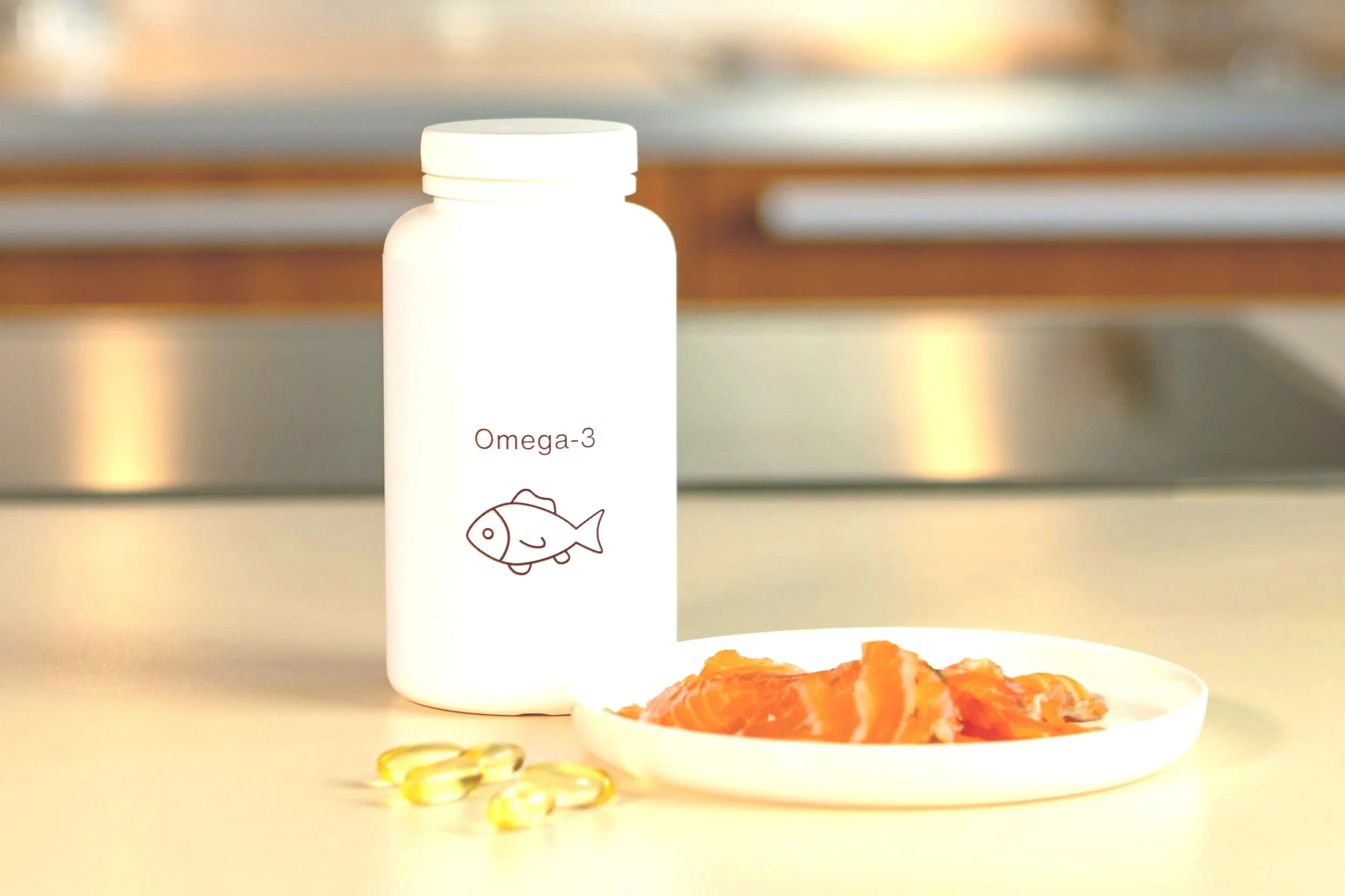 White supplement bottle labeled 'Omega-3' with an illustration of a fish, placed behind and to the left of a white plate with slices of smoked salmon, and some yellow fish oil capsules on the surface in front.
