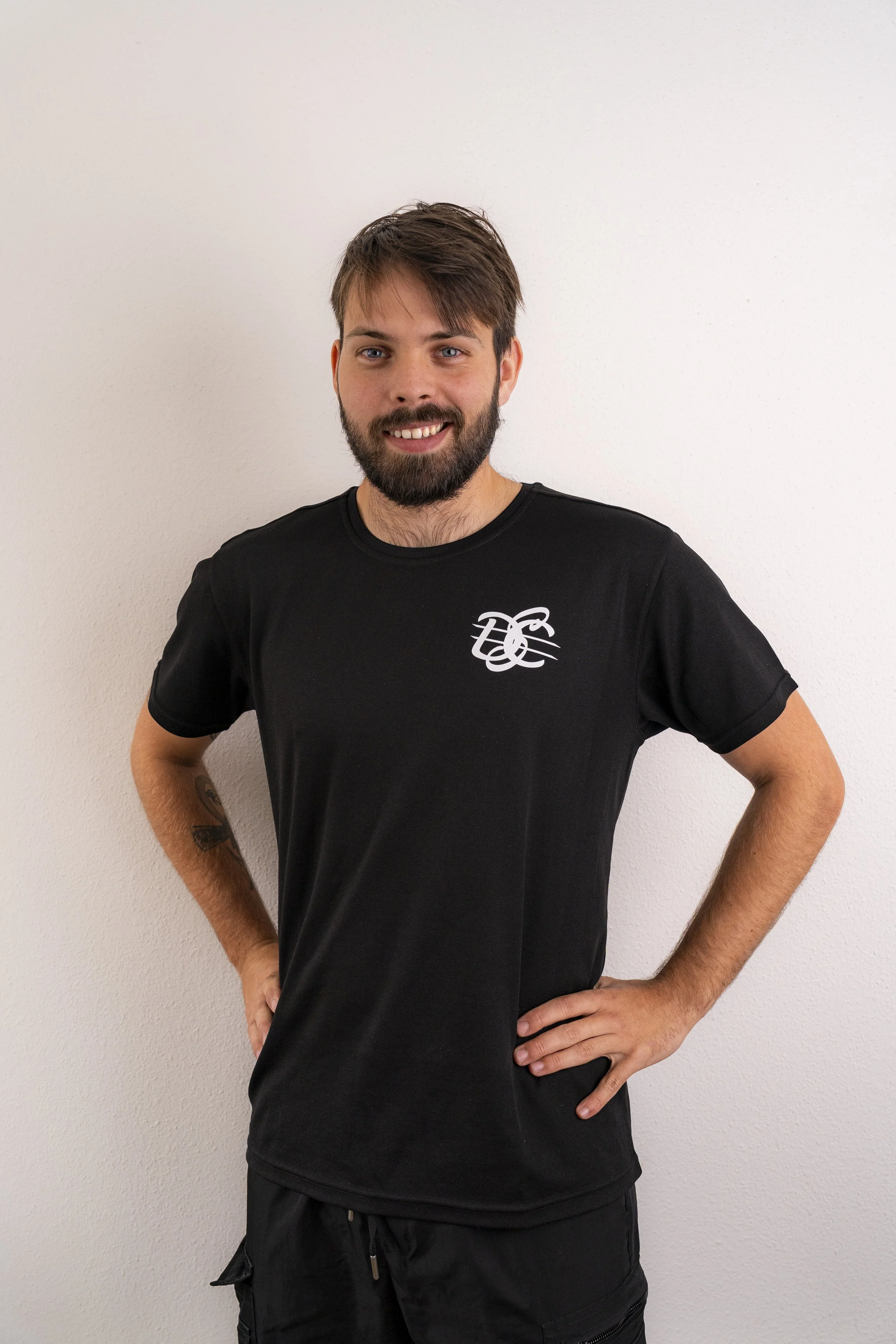 A smiling man with a beard and mustache standing with hands on hips, wearing a black t-shirt with a white logo on the chest, in front of a plain white wall.