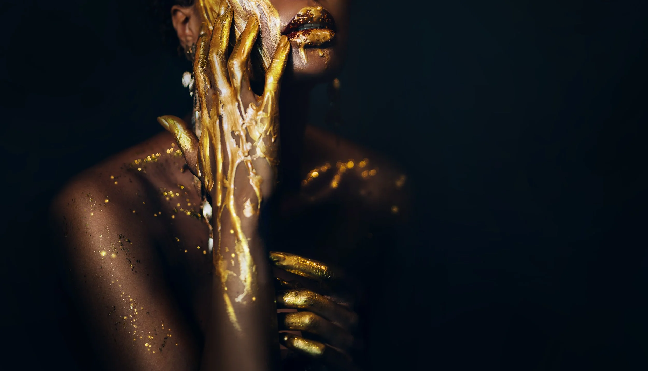 A woman with dark skin partially covered in gold paint or glitter, with her hand also painted gold, touching her face and shoulder against a dark background.