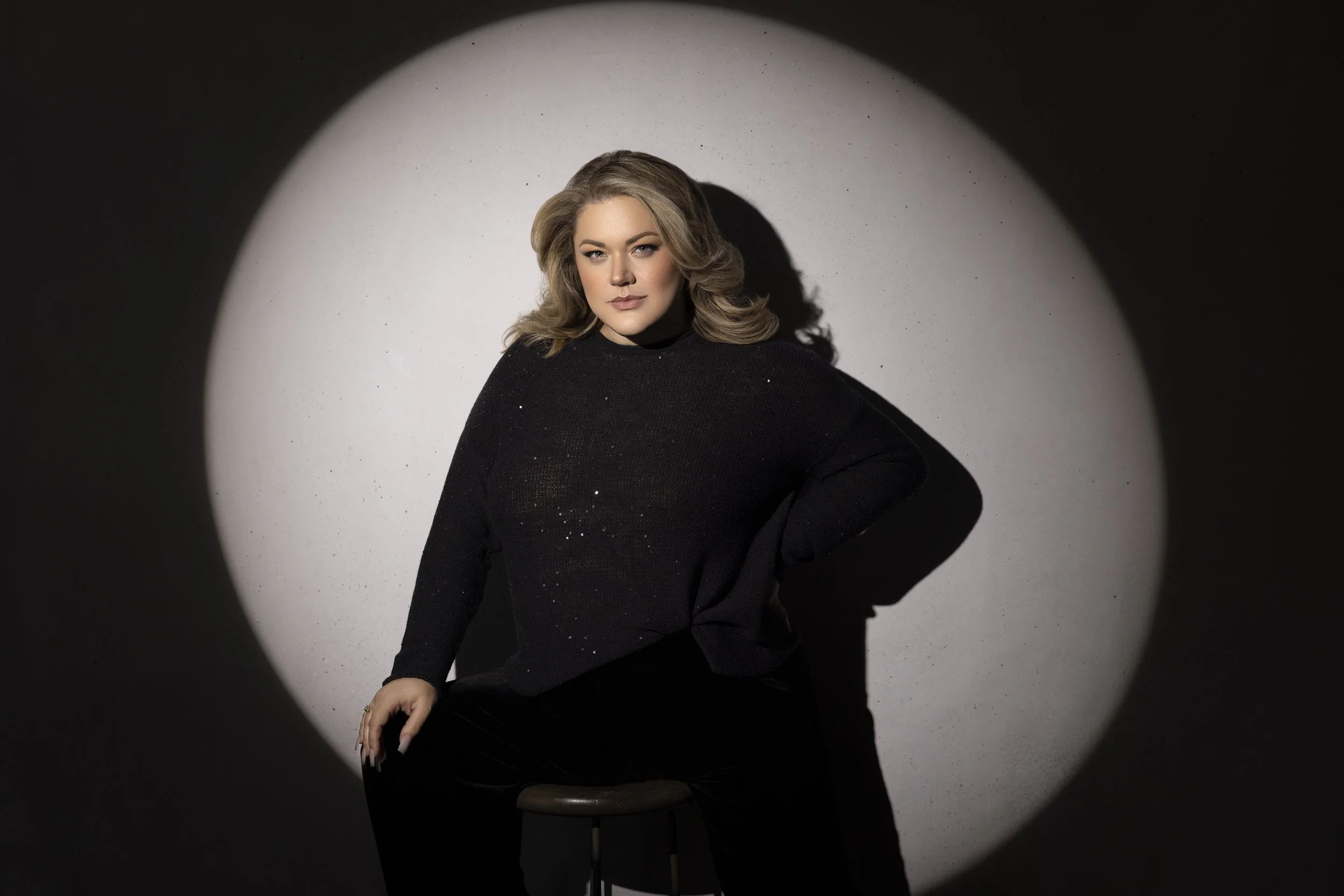 A woman with wavy blonde hair and makeup, wearing a black sweater, sitting on a stool, with a spotlight creating a halo effect behind her and a dark surrounding.