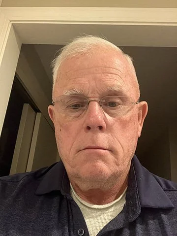 Older man with short white hair wearing glasses and a dark polo shirt, standing indoors.