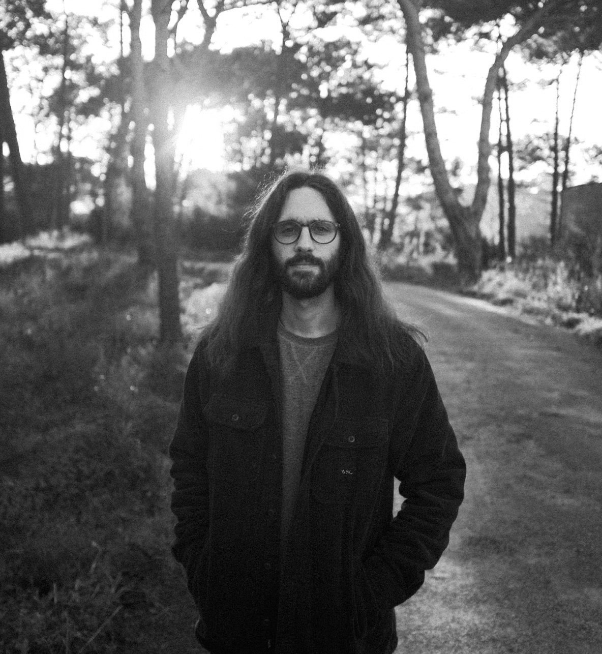 A man with long hair, glasses, and a beard standing outdoors on a dirt path surrounded by trees.