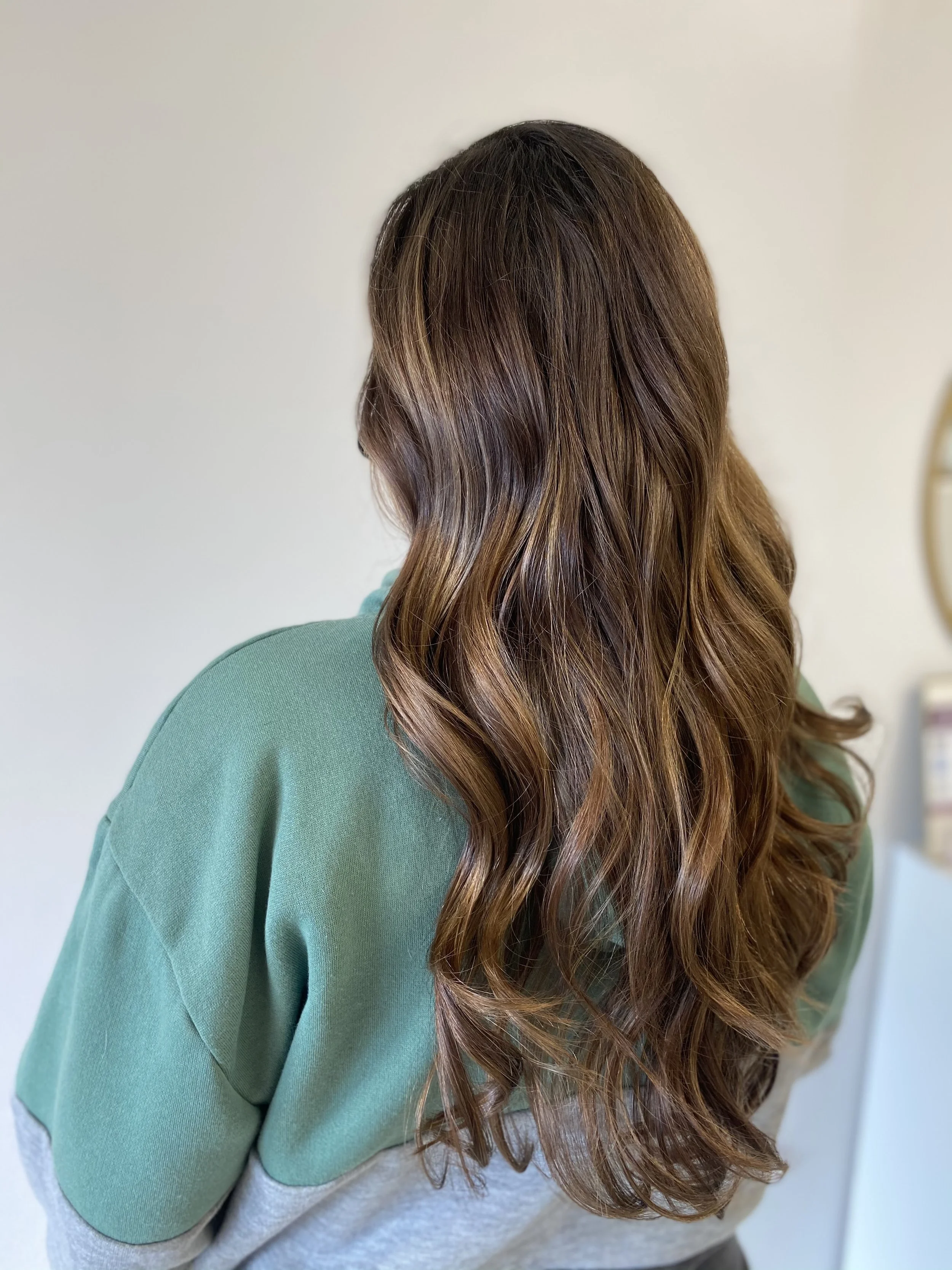 Back view of a woman with long, wavy brown hair wearing a green sweatshirt.