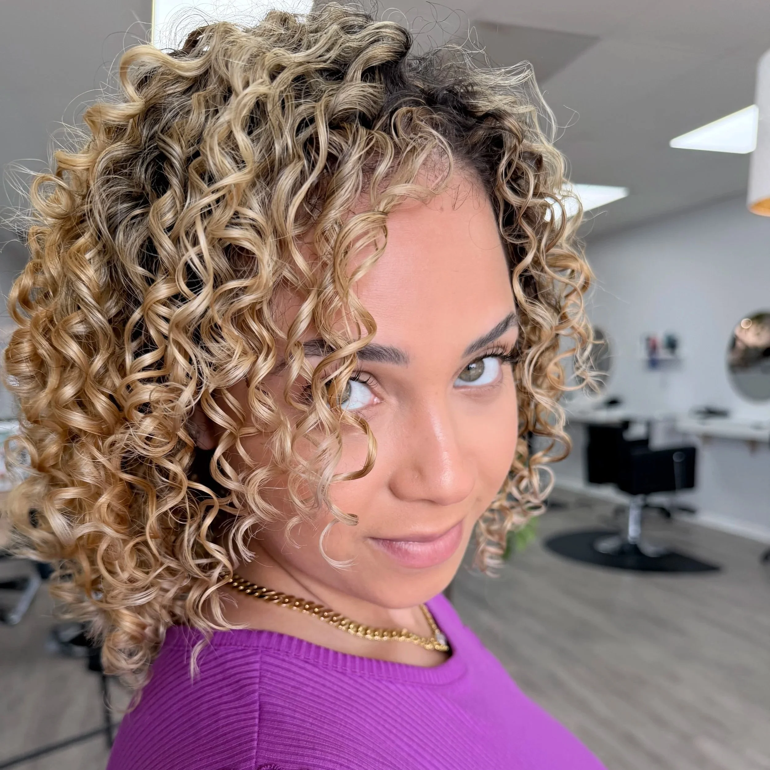A woman with curly blonde hair wearing glasses, layered gold necklaces, and an off-the-shoulder gray t-shirt, standing indoors near a plant and decorative hanging lights.