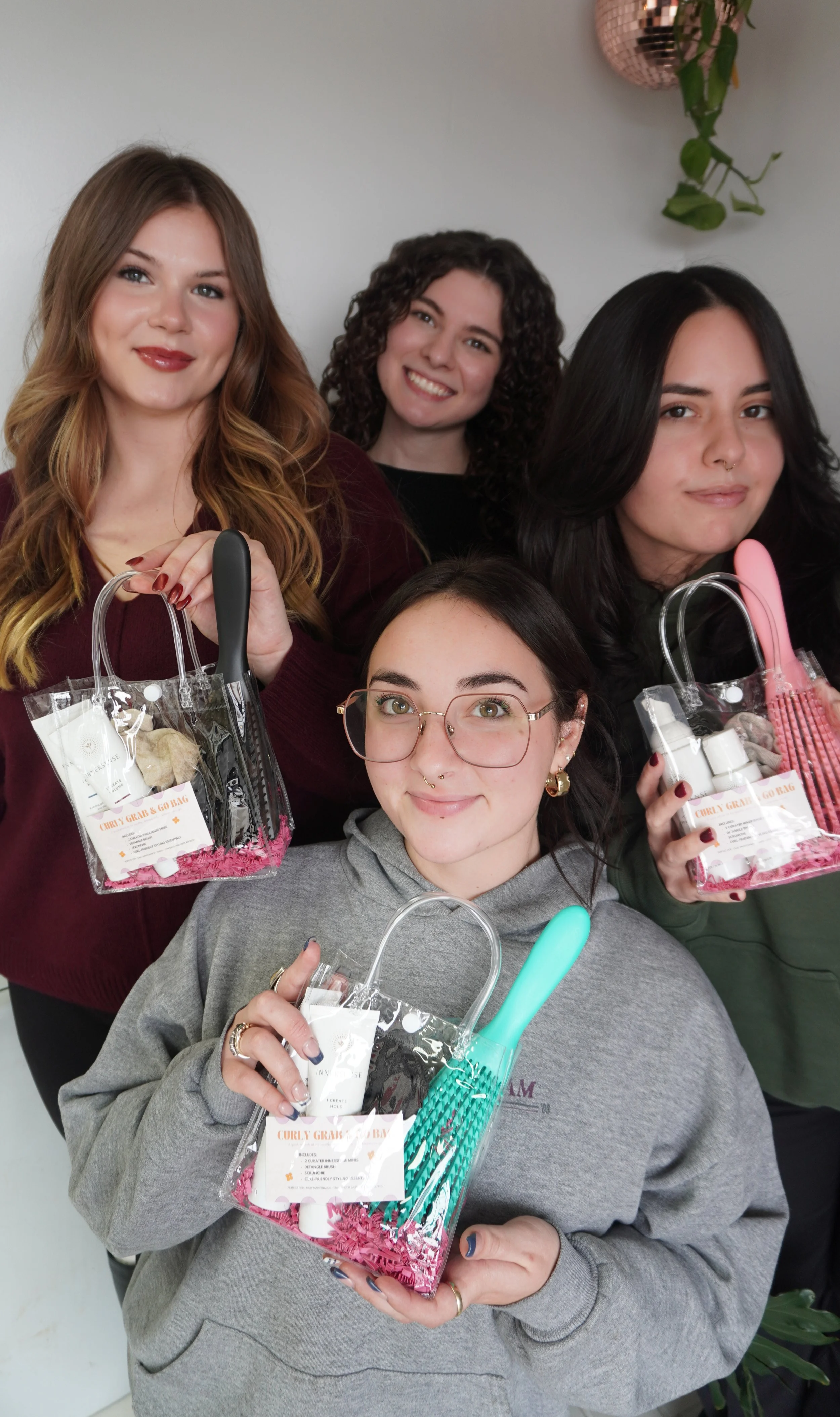 A group of five women holding small gift bags with beauty products, brushes, and toiletries, smiling indoors against a light-colored wall.