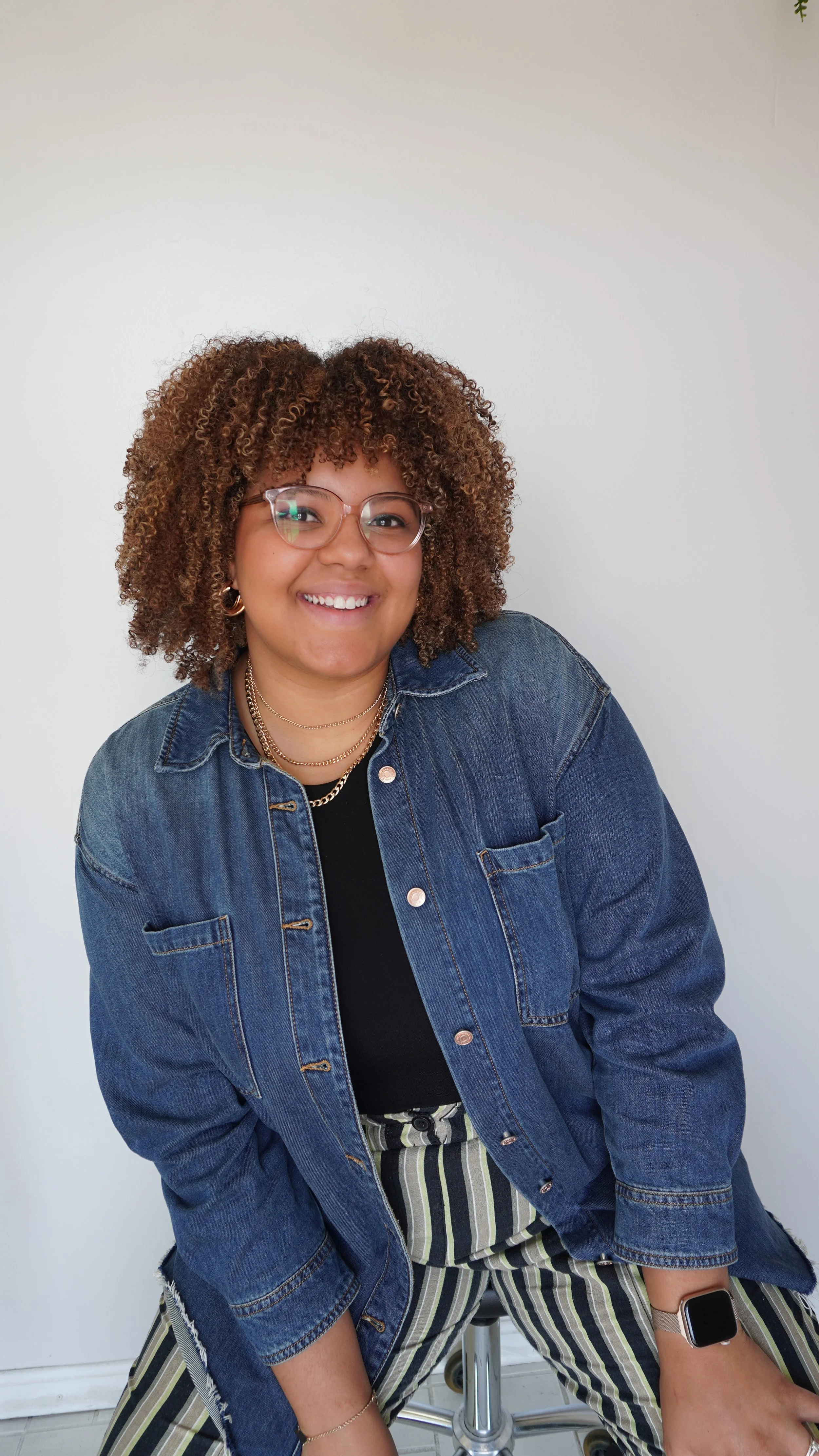 A smiling woman with curly brown hair, wearing glasses, a yellow top, and a dark denim jacket, standing in front of a plain light-colored wall with a plant in the background.