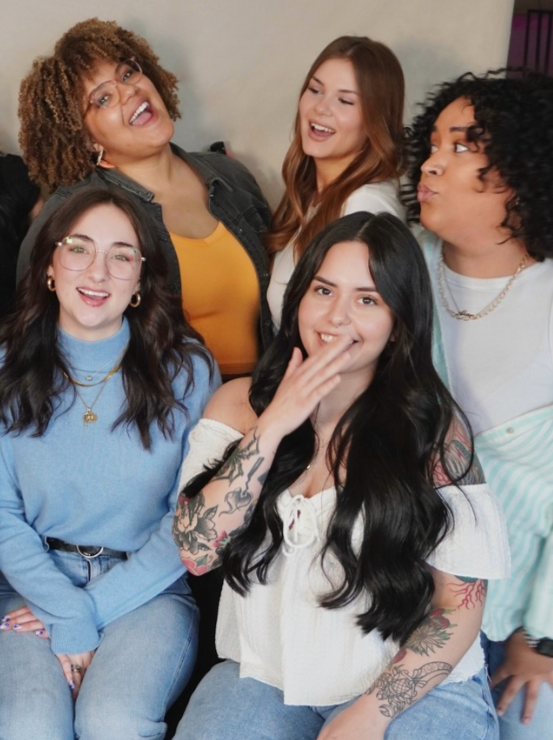 Group of five diverse women smiling and laughing together, some making playful expressions, in a casual indoor setting.