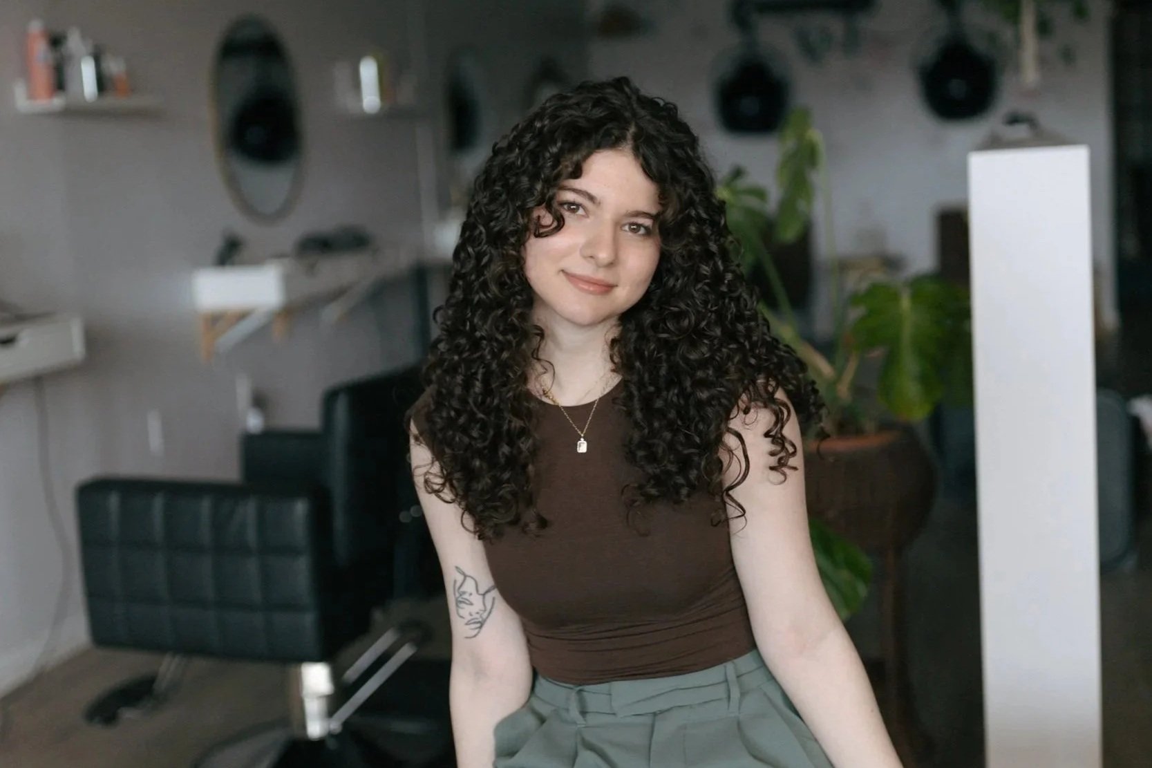 A smiling woman with curly hair and glasses, wearing a purple corduroy jacket and jewelry, standing against a white wall with a pink disco ball planter and green plant above her.