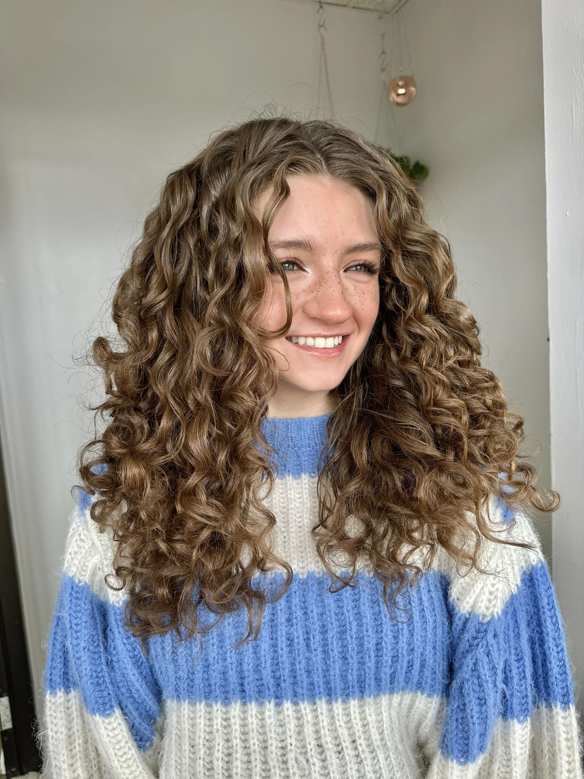 A young woman with long, curly brown hair and freckles smiling while wearing a blue and white striped sweater in an indoor setting.