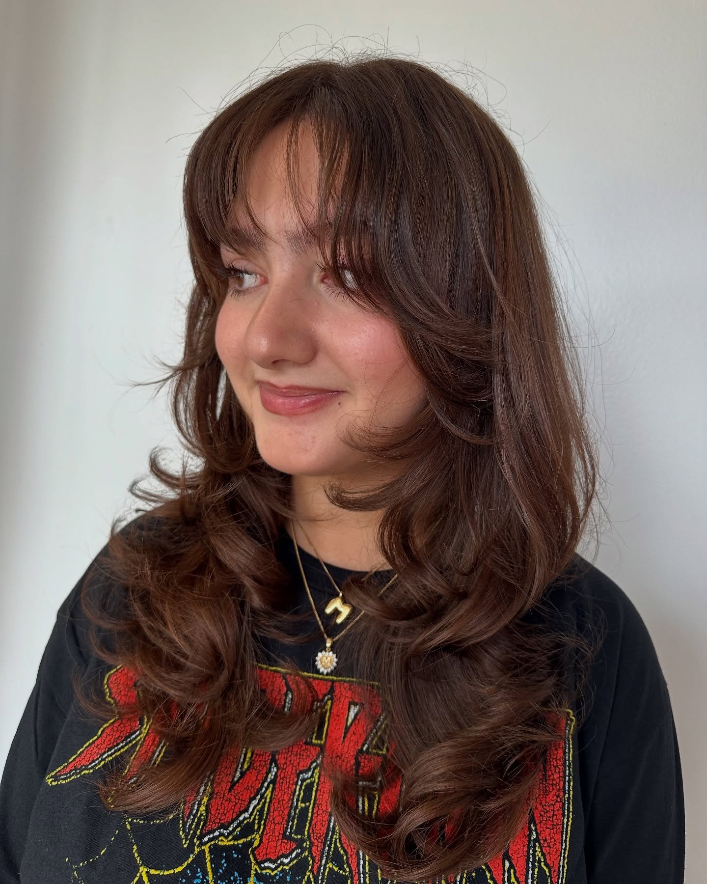 Young woman with wavy brown hair wearing a black T-shirt with red and yellow text and layered gold necklaces, posing against a plain light-colored wall.