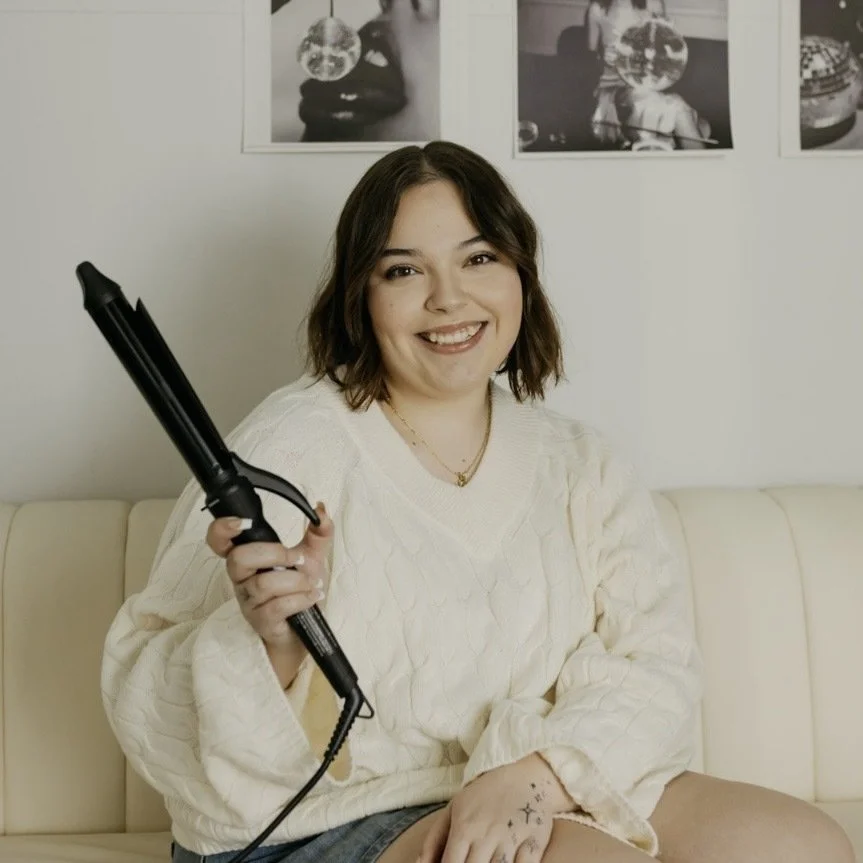 A young woman with long brown hair and light skin, smiling and sitting on a beige sofa with a large green leafy plant behind her, wearing a black long-sleeve top, a denim vest, and layered gold necklaces.