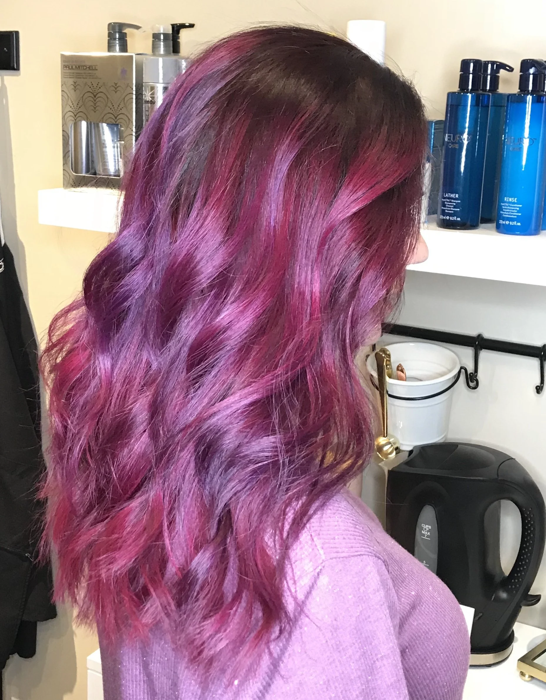Woman with wavy hair dyed in pink and purple shades, wearing a pink top, standing in a hair salon with hair care products on shelves behind her.