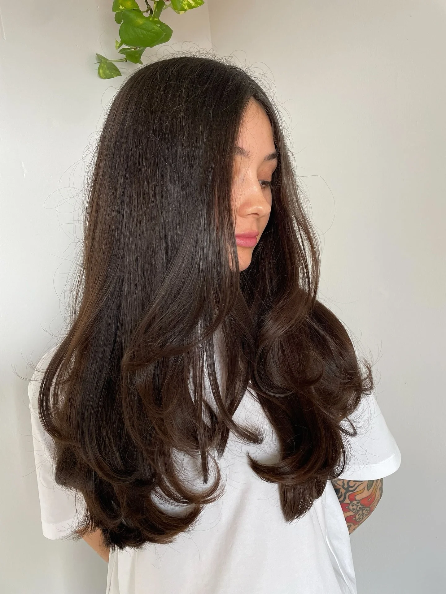 Side view of a woman with long, dark brown, wavy hair standing against a plain wall with a small green plant nearby.