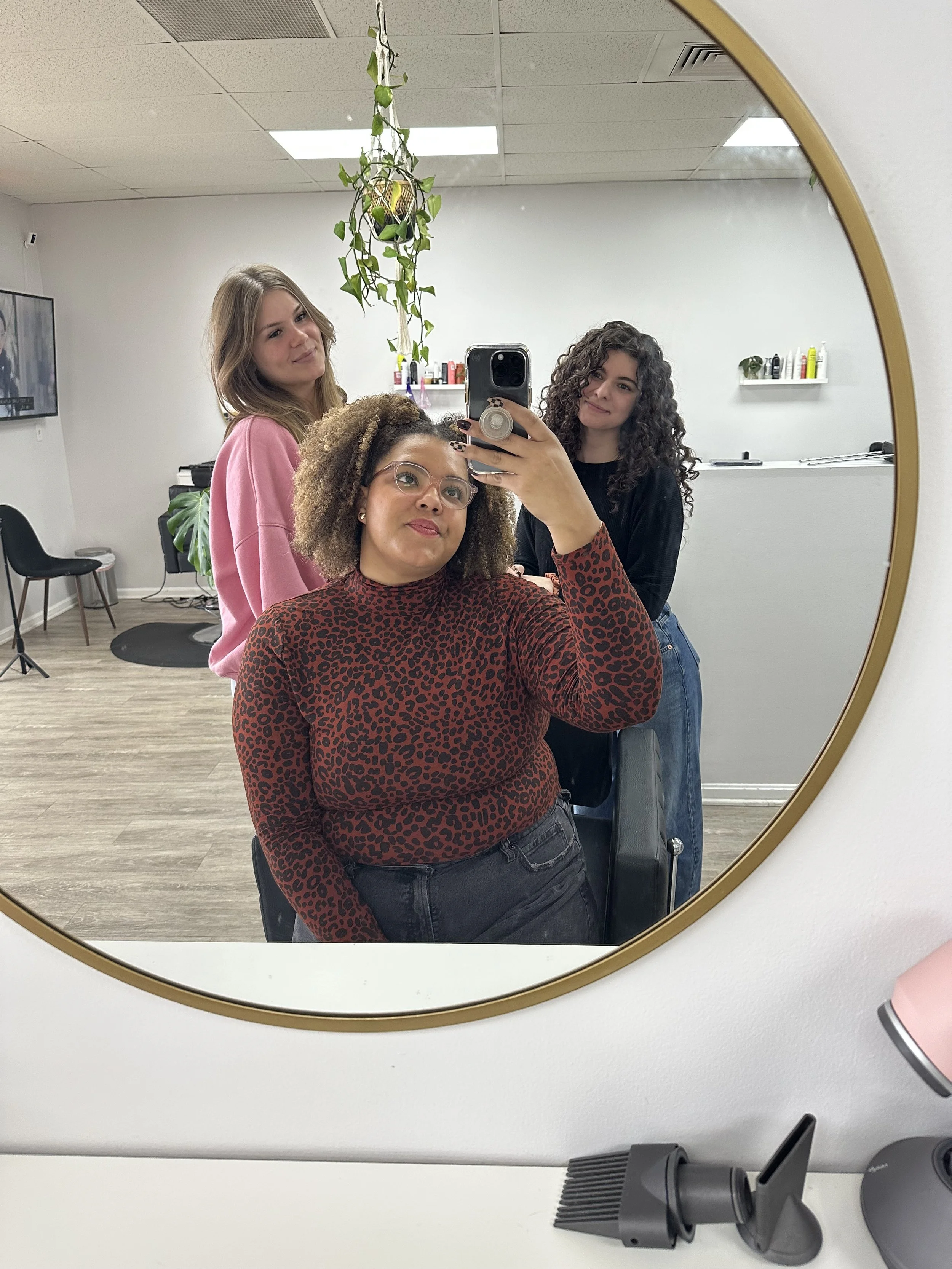 Three women with different hair types and skin colors taking a selfie in a hair salon mirror, with one seated and two standing behind her, all smiling.
