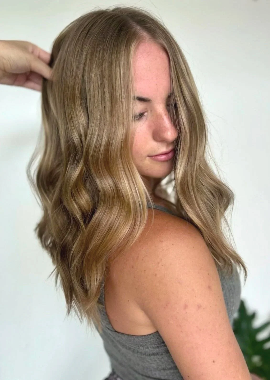 A woman with long, wavy blonde hair, wearing a gray sleeveless top, is getting her hair styled by someone whose hand is visible adjusting her hair. The background is plain and light-colored.
