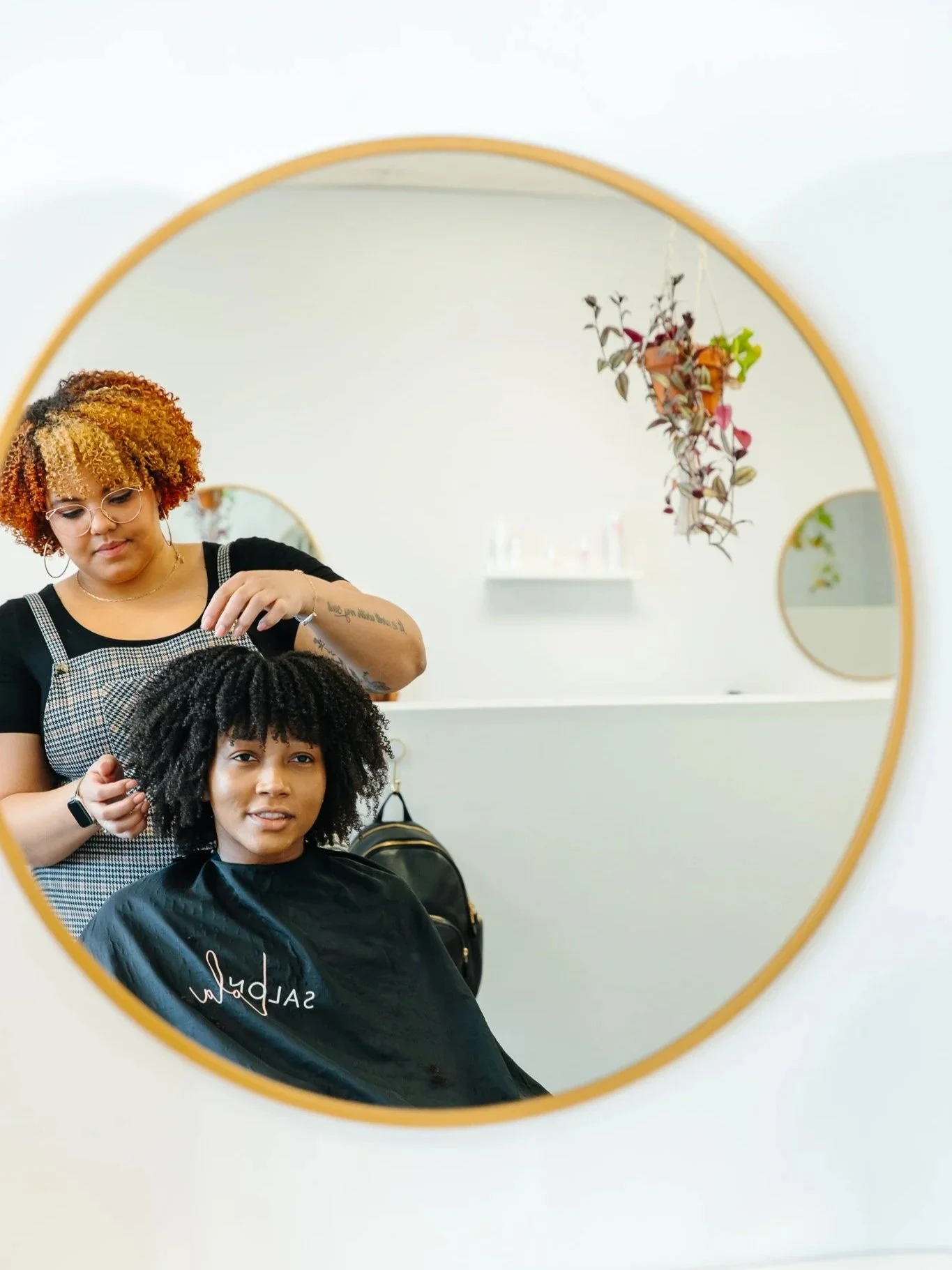 Hairdresser styling a woman's curly hair in front of a round mirror in a salon.